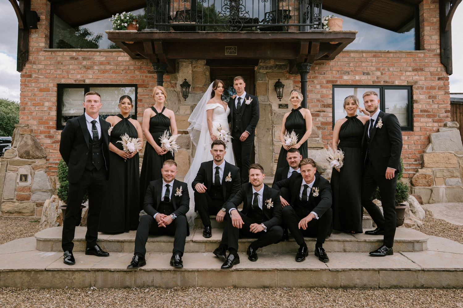 Wedding party in black formal attire on stone steps in front of brick building on wedding day. Wedding photography at Willow Marsh Farm.