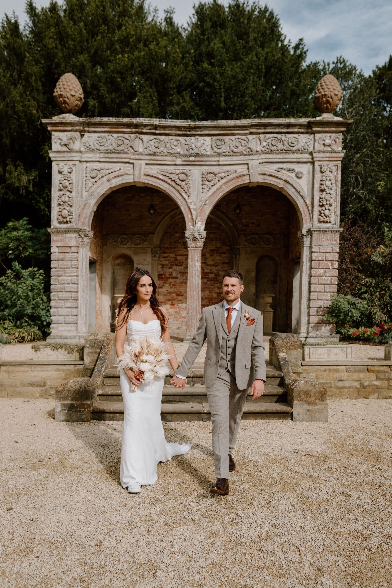 Couple standing in front of an ornate stone archway in a garden setting.