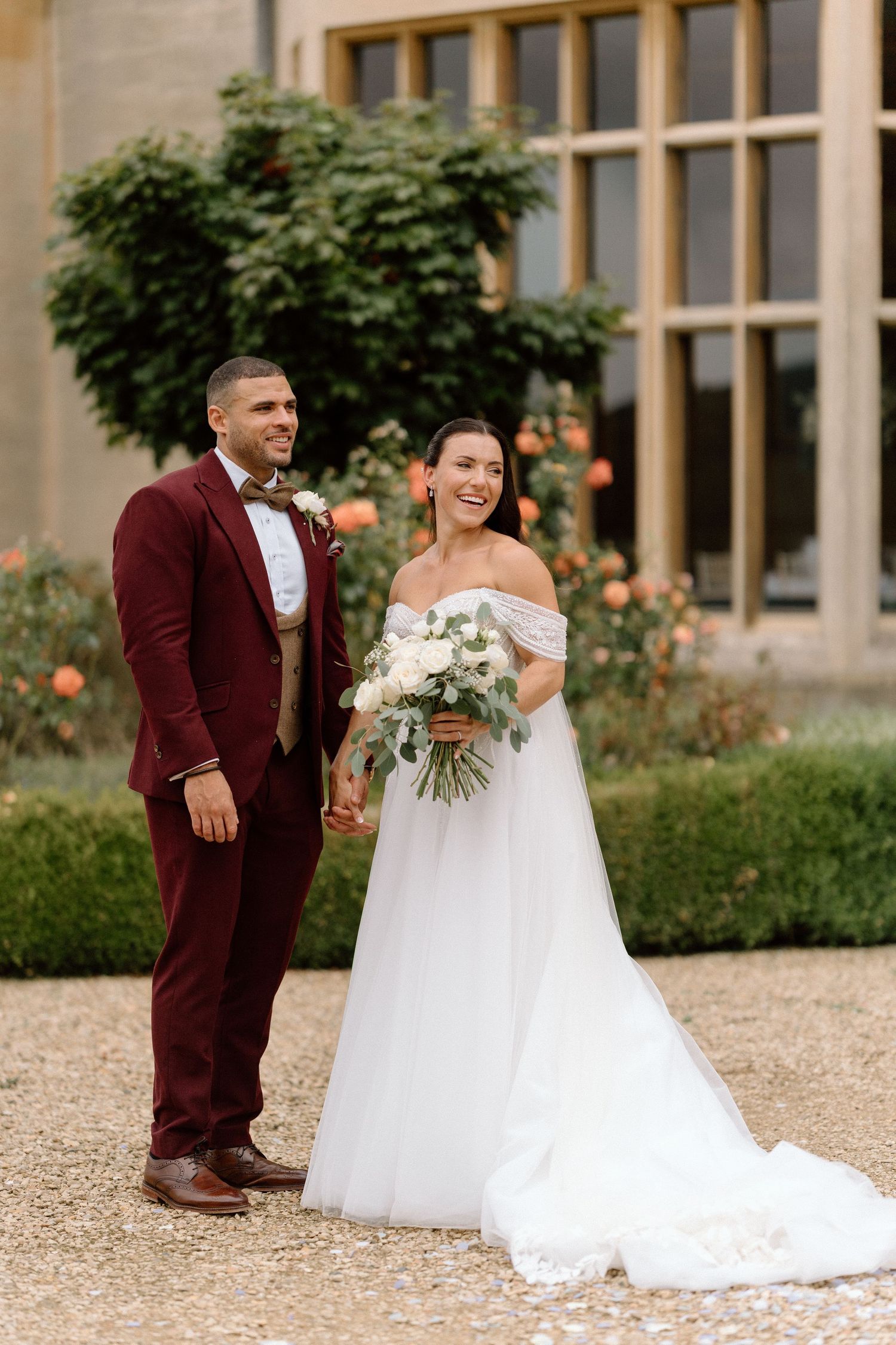 Wedding couple in a garden with groom in burgundy suit and bride in off-shoulder white gown holding floral bouquet at Harlaxton Manor.