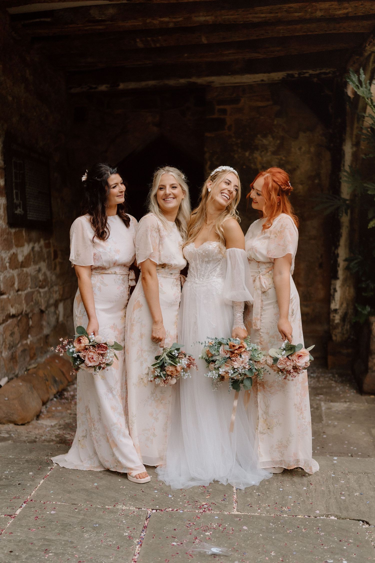 Four bridesmaids in light colored dresses pose together holding floral bouquets against a stone wall backdrop Cheylesmore Manor in Coventry.