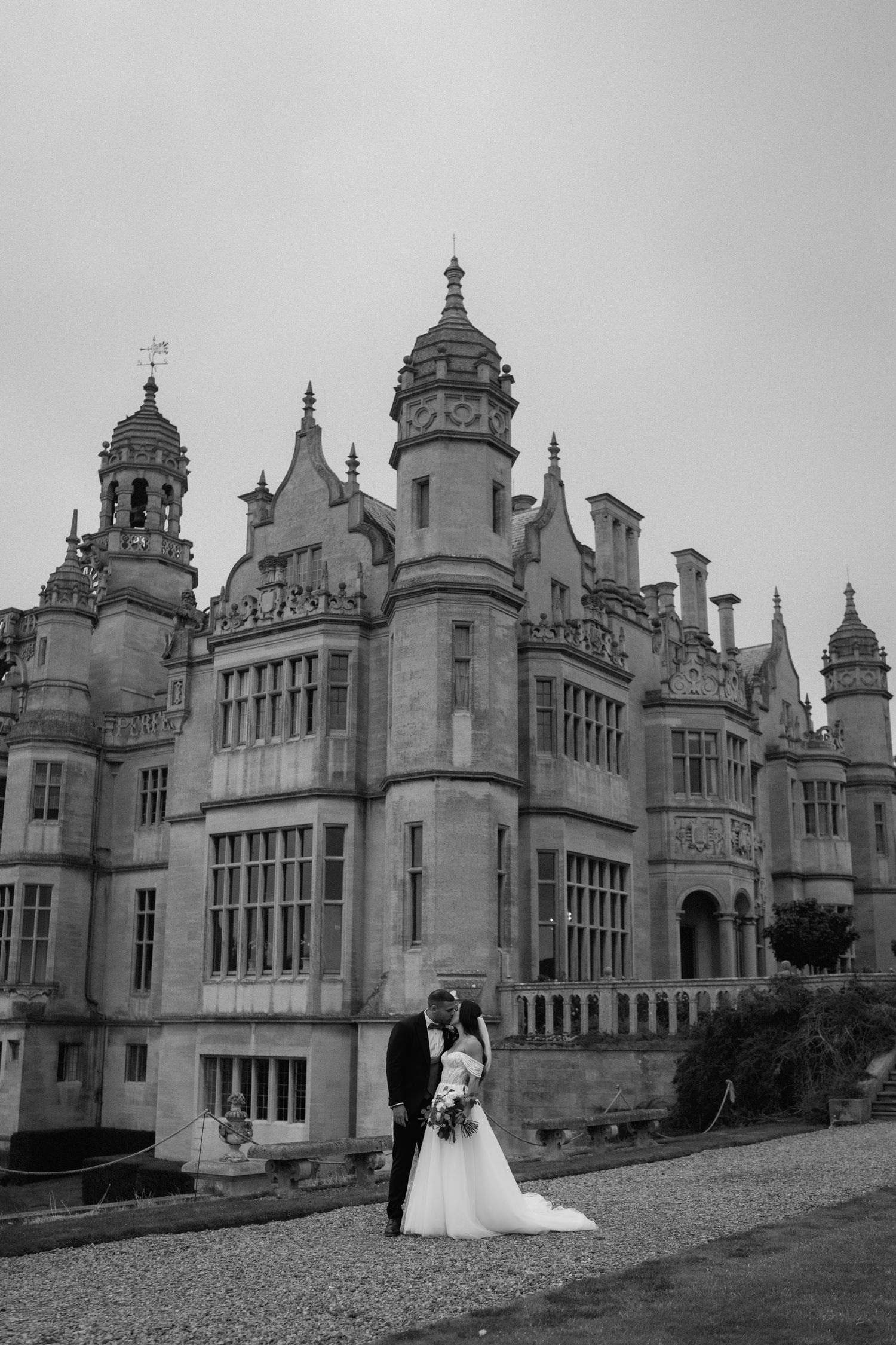 Black and white photo of newlyweds in front of a grand Gothic-style castle with ornate architecture at Harlaxton Manor.