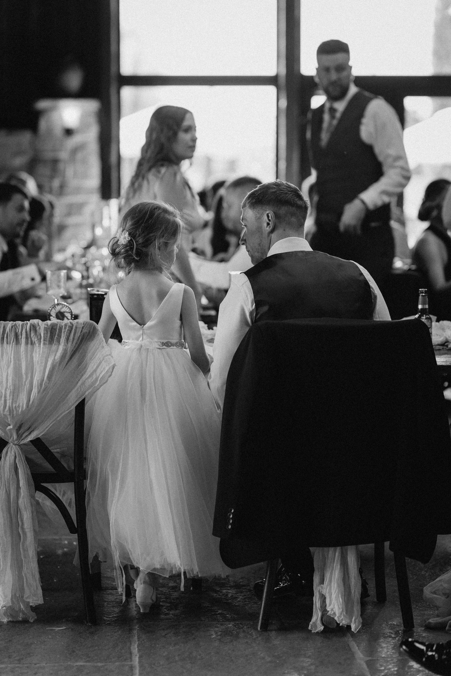 A tender black and white wedding reception moment as guests gather at tables in warm indoor venue. Wedding photography at Willow Marsh Farm.