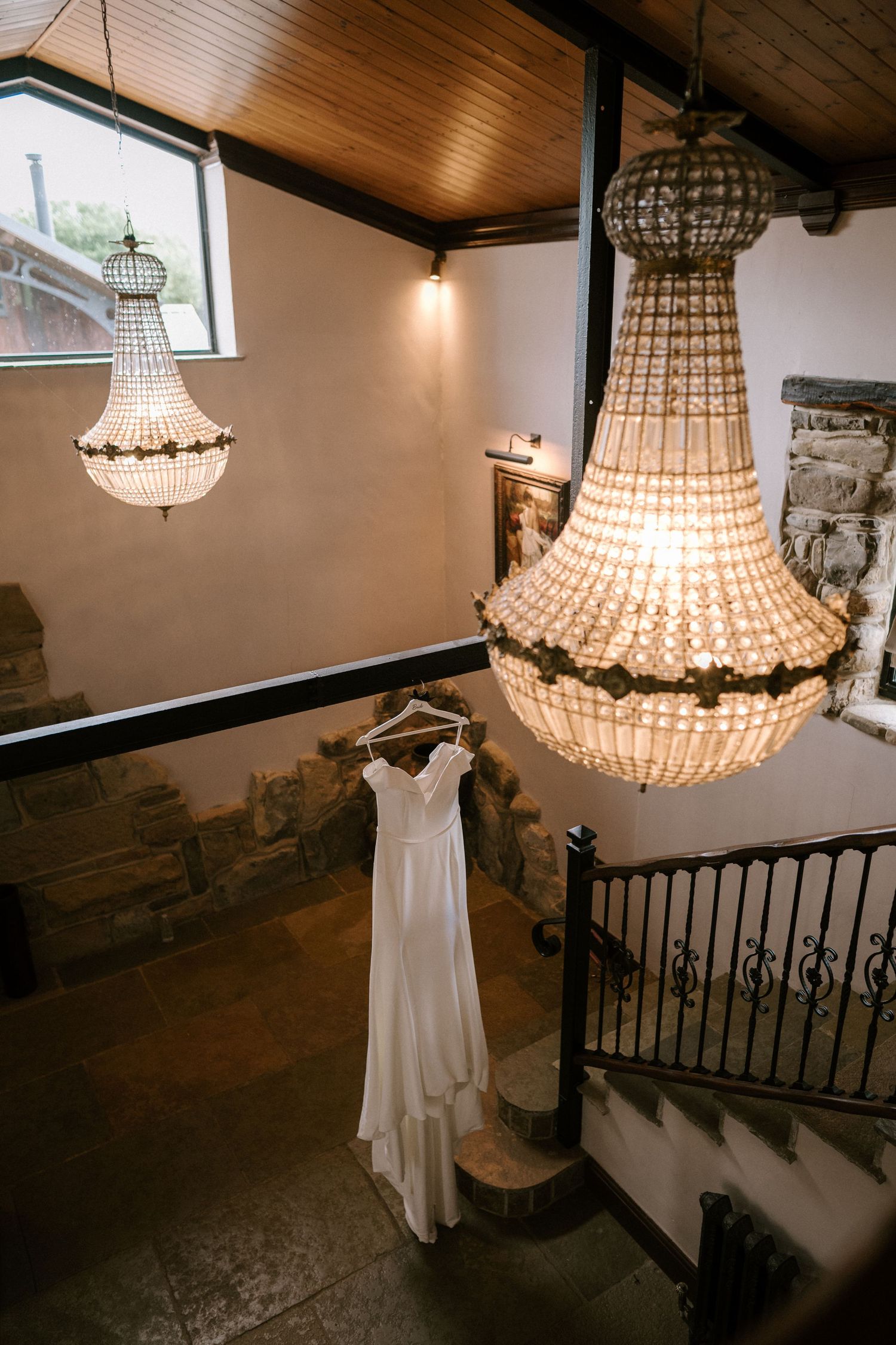 Elegant white wedding dress hanging beneath crystal chandeliers in rustic stone stairwell. Wedding photography at Willow Marsh Farm.