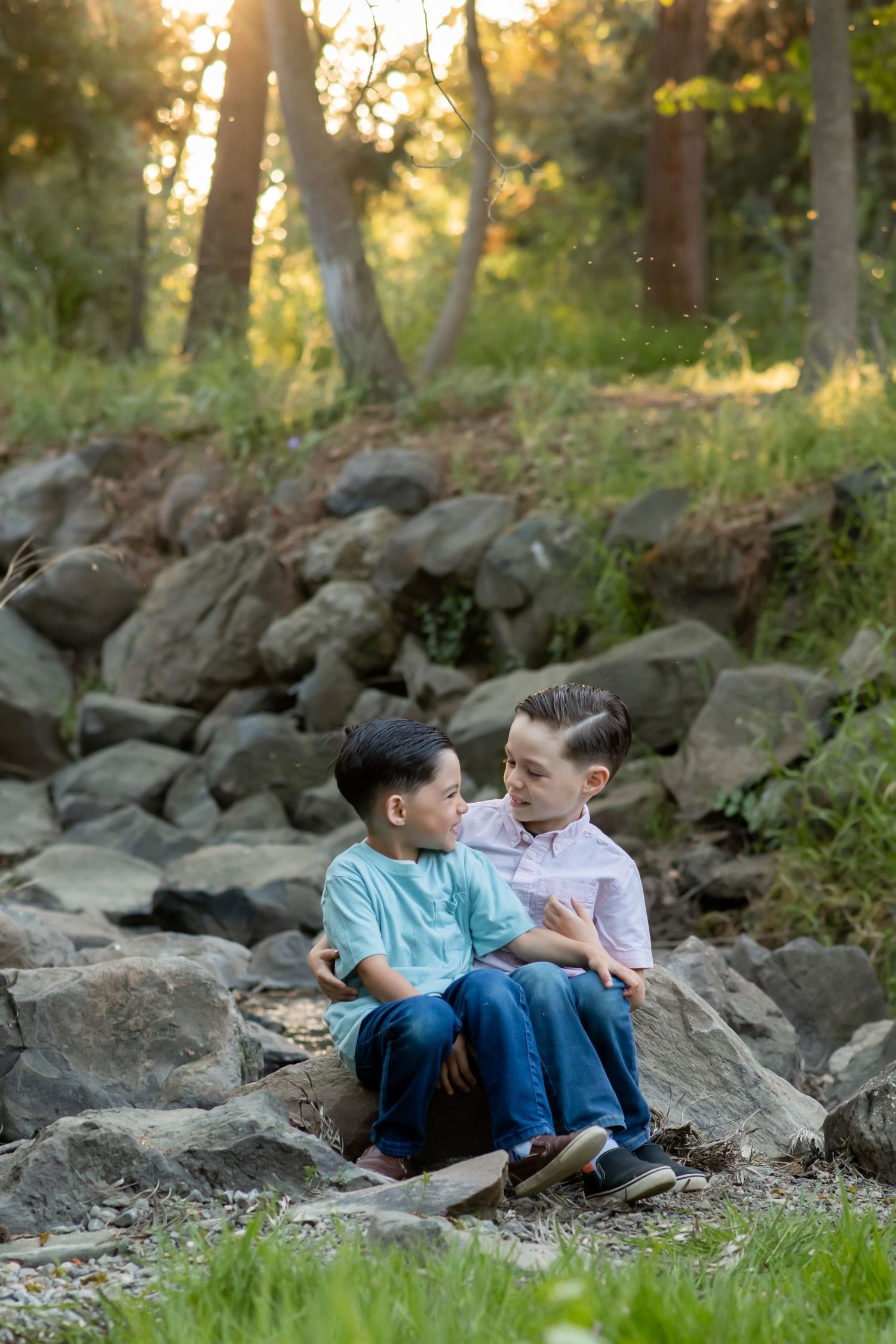 Two young boys sit together on large rocks in a woodsy outdoor setting during golden hour.