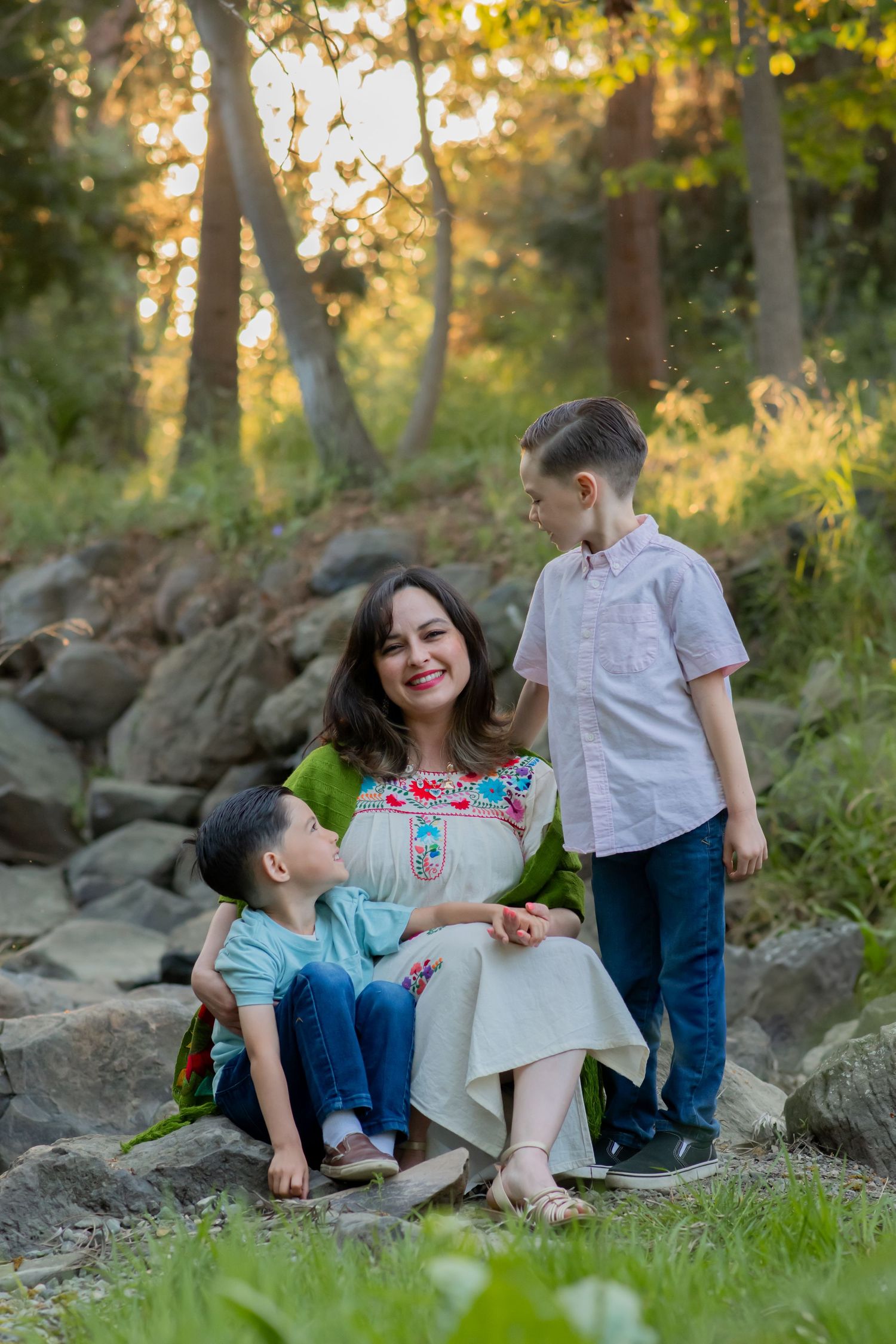 A happy family shares a sweet moment together on rocky terrain during golden hour in a sunlit forest setting.