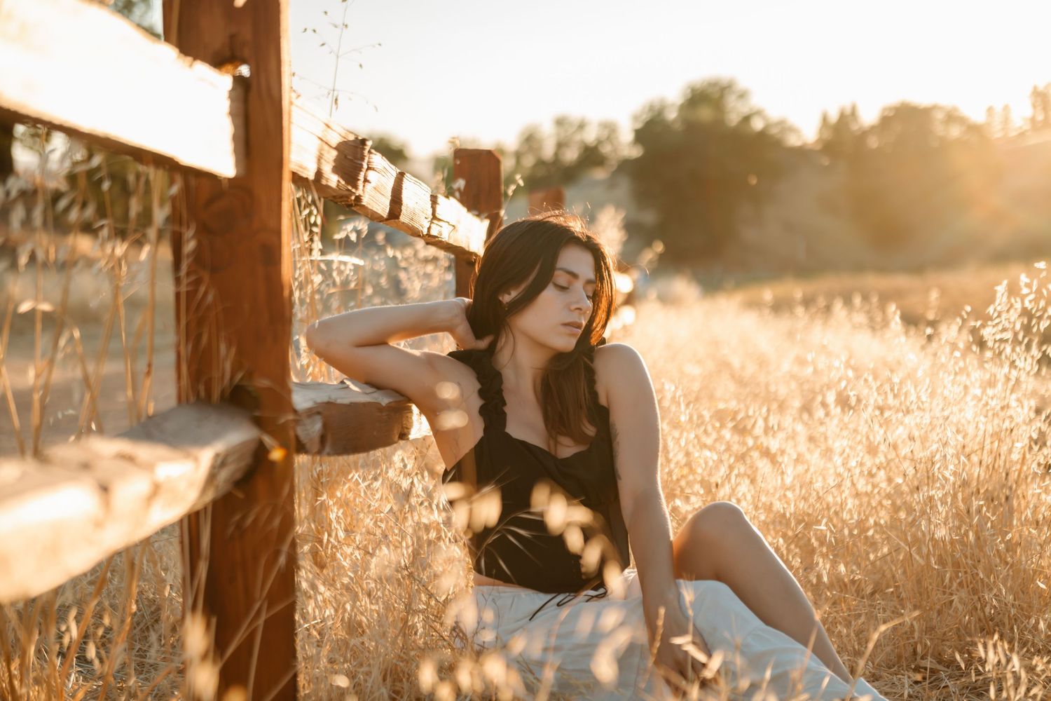 A person sits in a golden field at sunset while wearing a black dress and denim.