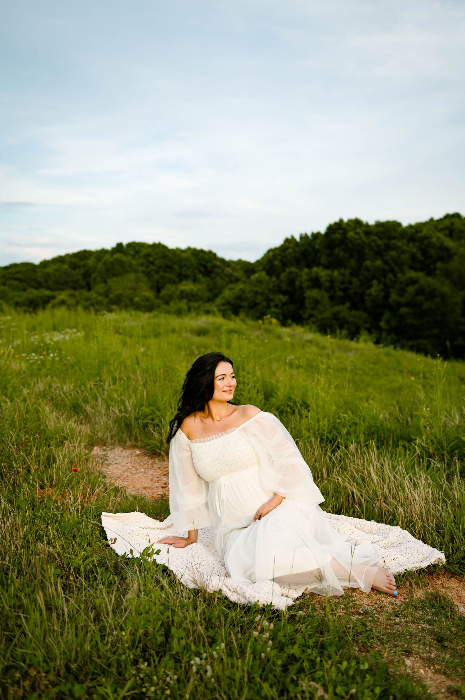 Mother in a flowing white dress sits gracefully in a green meadow with wooded hills in the background with durham maternity photographer