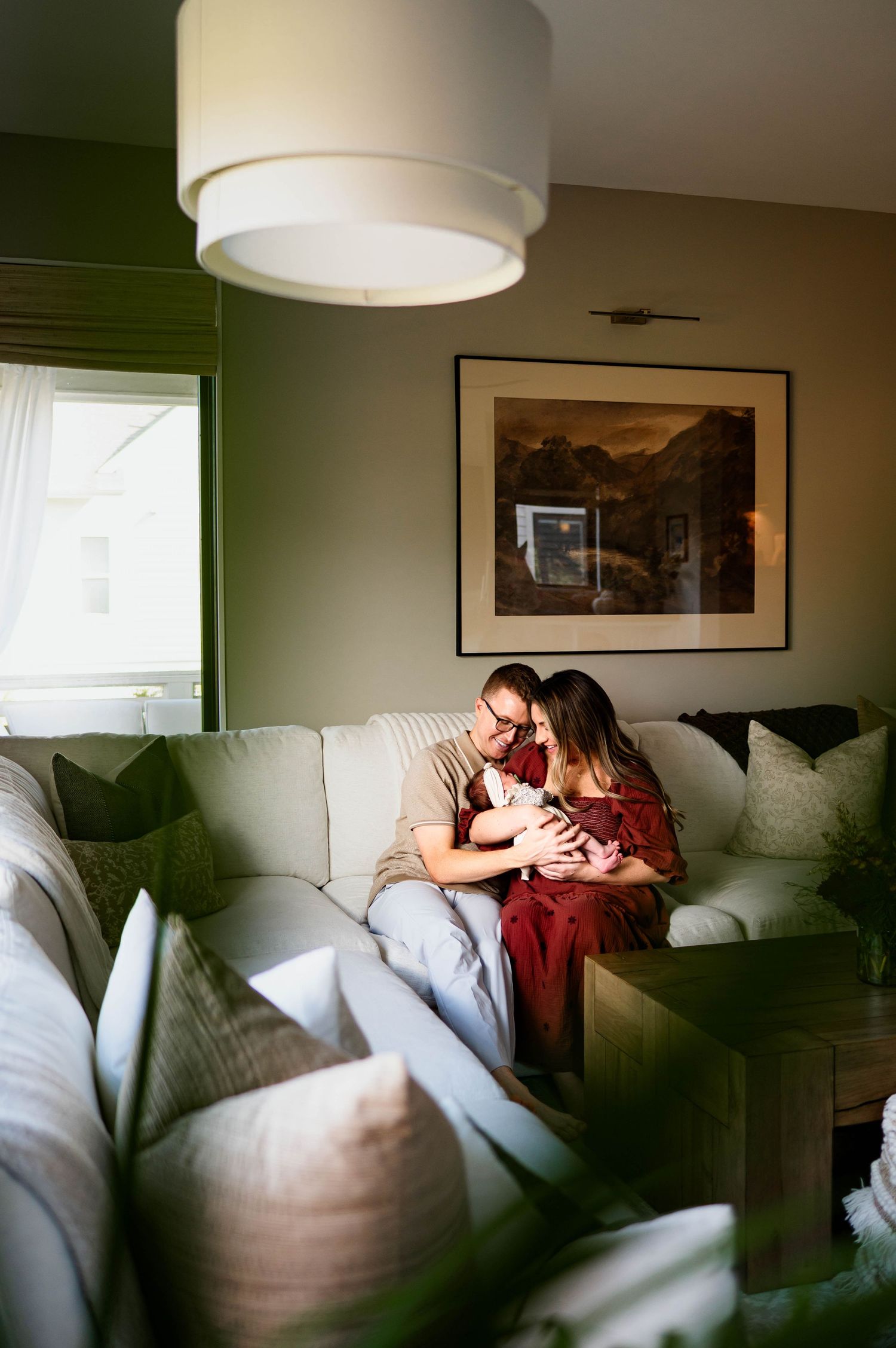 A family relaxes together on a plush white couch in a cozy living room with green walls with durham newborn photographer