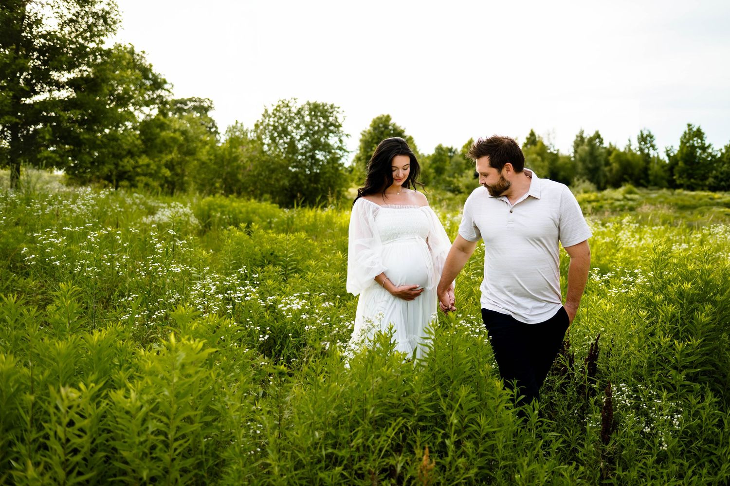 Expecting couple walks hand in hand through a lush wildflower meadow on a sunny summer day with raleigh maternity photographer