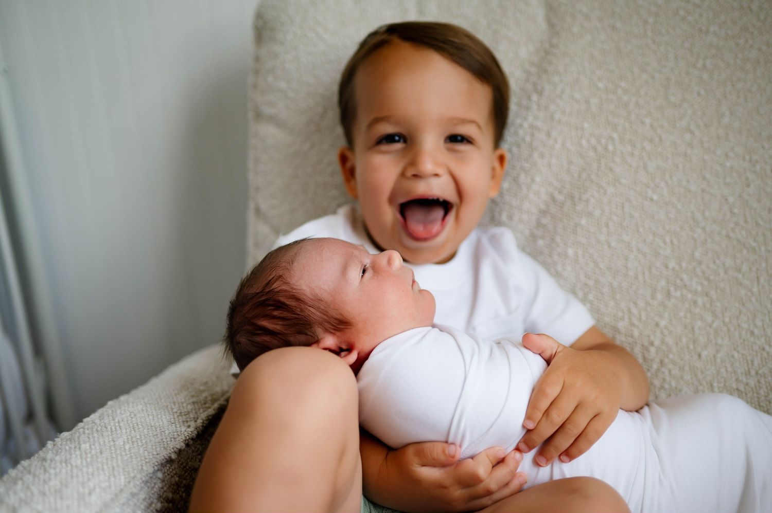 Excited brother holds his newborn brother while sitting on a light colored chair and looks at camera for photo by best newborn photographer