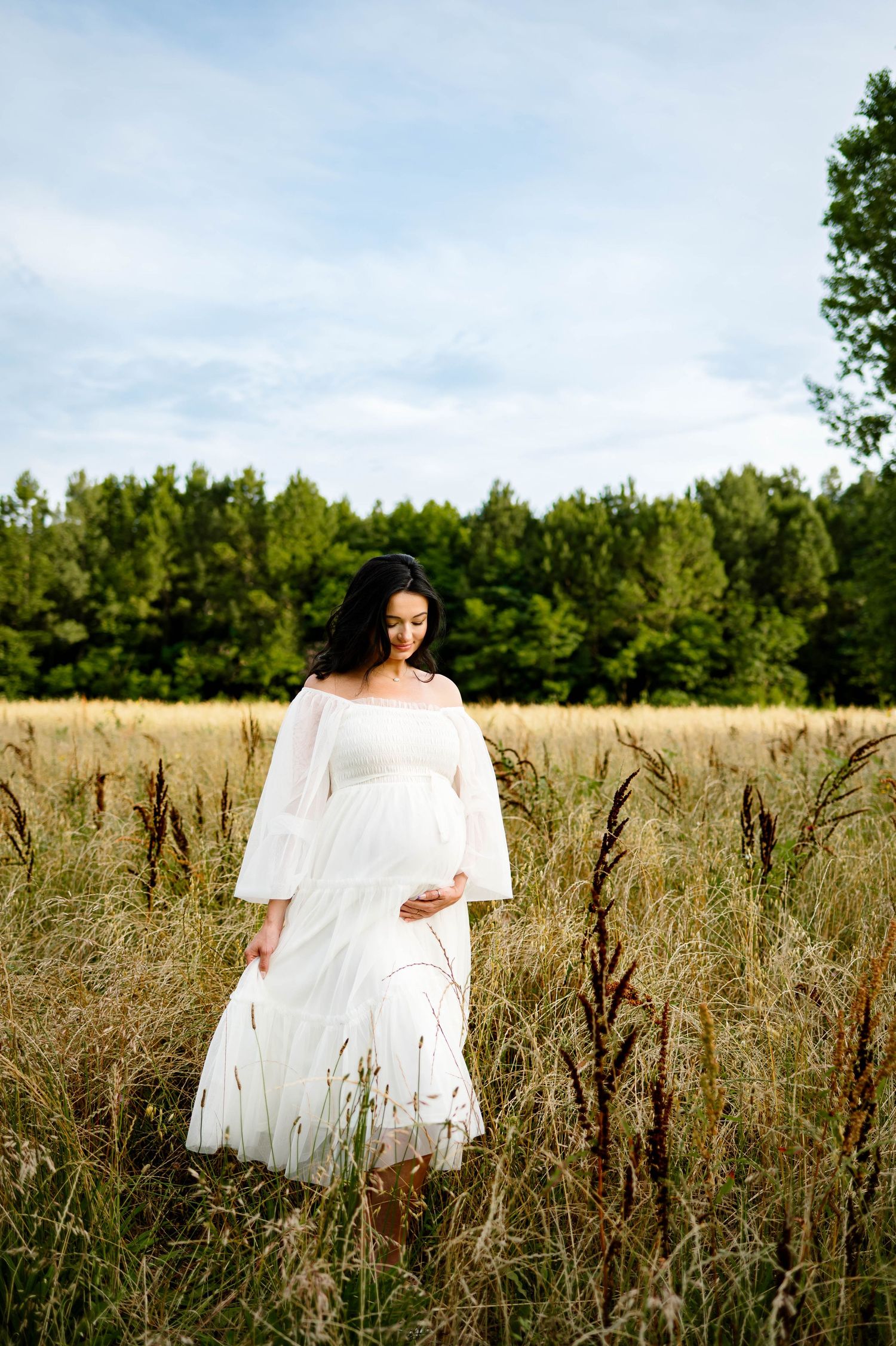 A mother in a white dress stands among tall grass in a field at sunset in image from maternity photography session in raleigh