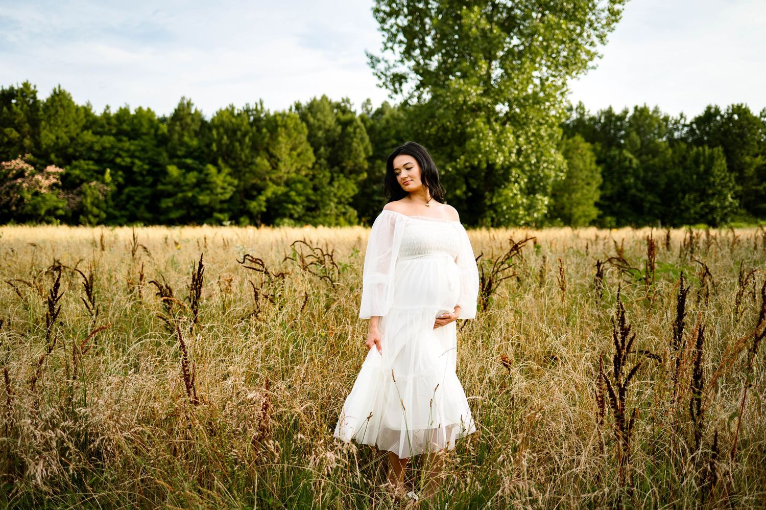 Mother in a flowing white dress stands in a sun-lit meadow surrounded by tall grass and trees for image by raleigh maternity photographer