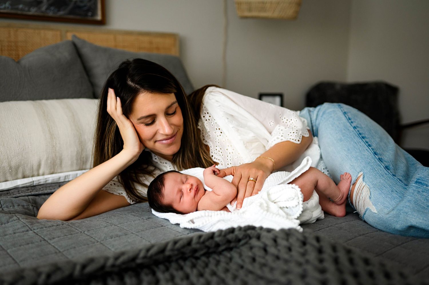 Mother relaxes on a cozy gray bed, sharing a tender moment with newborn in natural lighting during newborn photography session apex