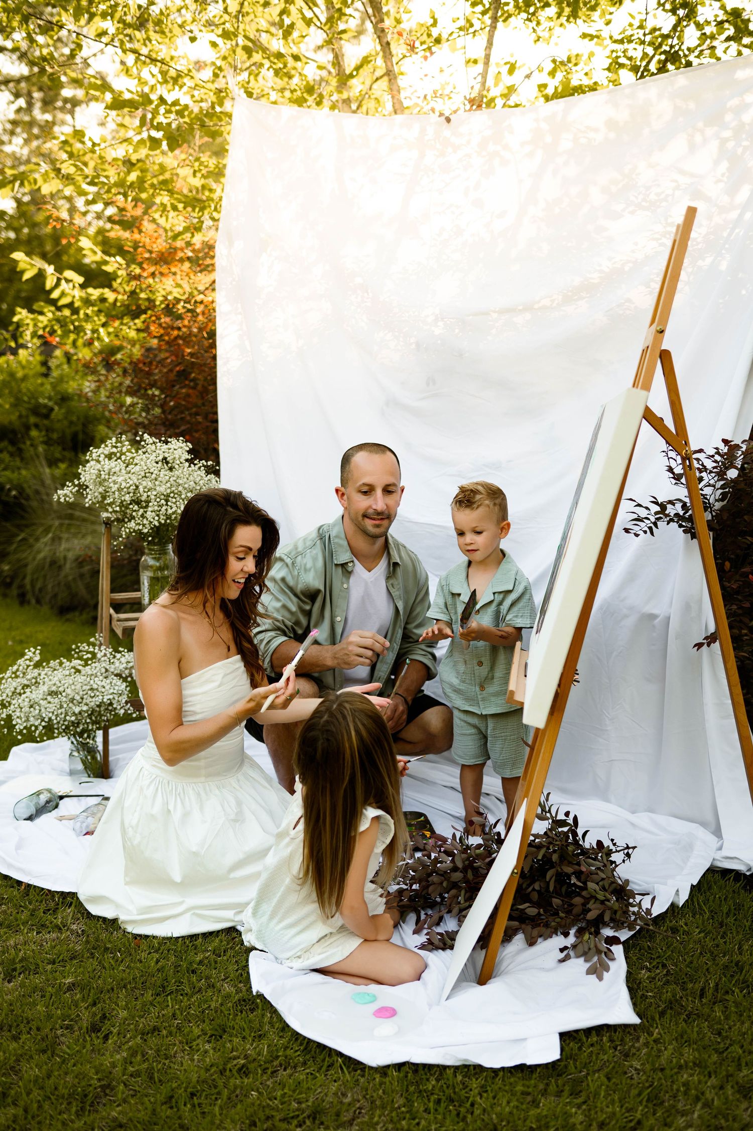 Family sitting on white blankets outdoors painting together during a summer photoshoot with best raleigh family photographer