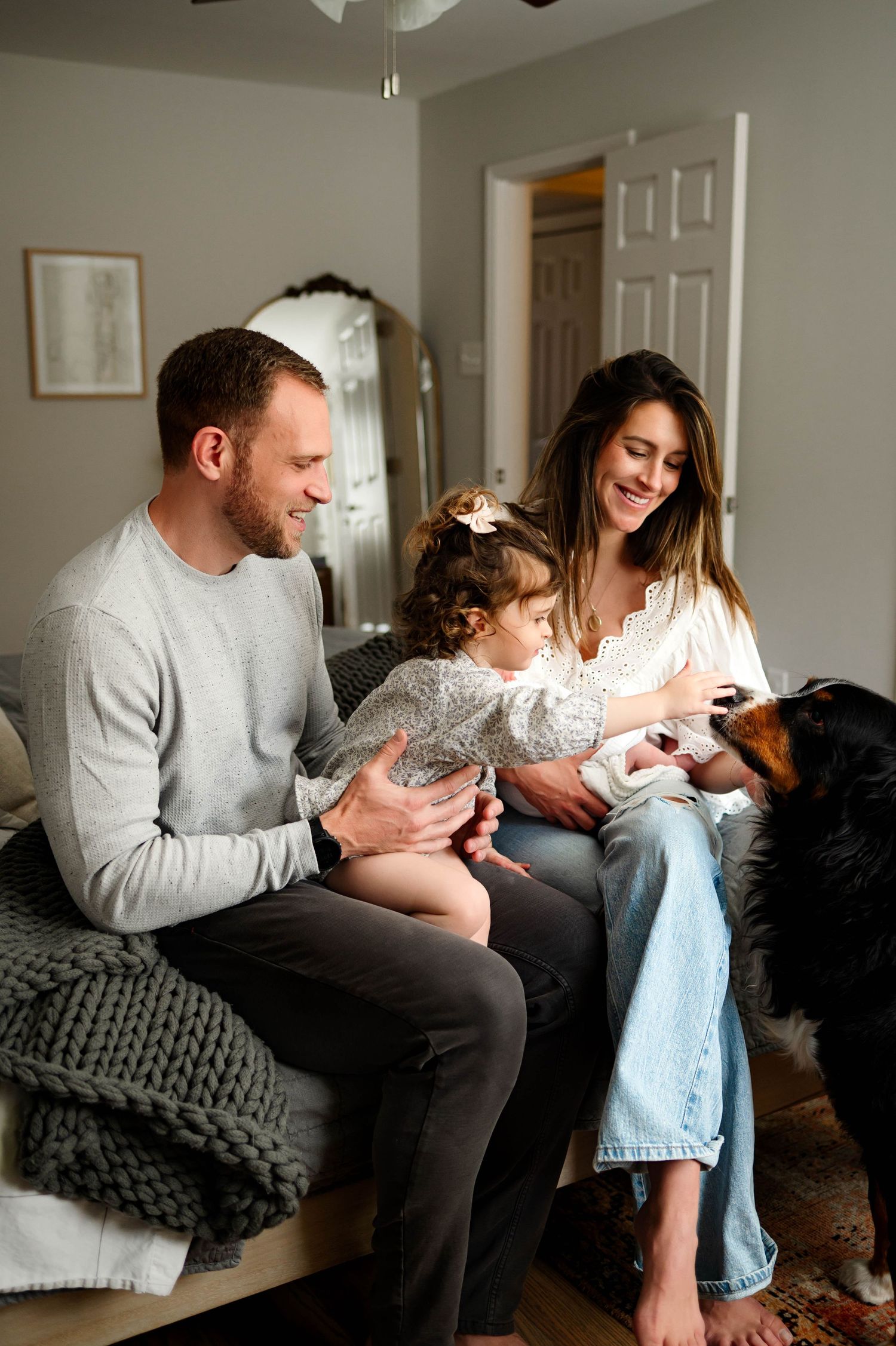 A family sits together on a bed in a cozy bedroom with their dog and newborn baby while enjoying their in home newborn photoshoot cary nc