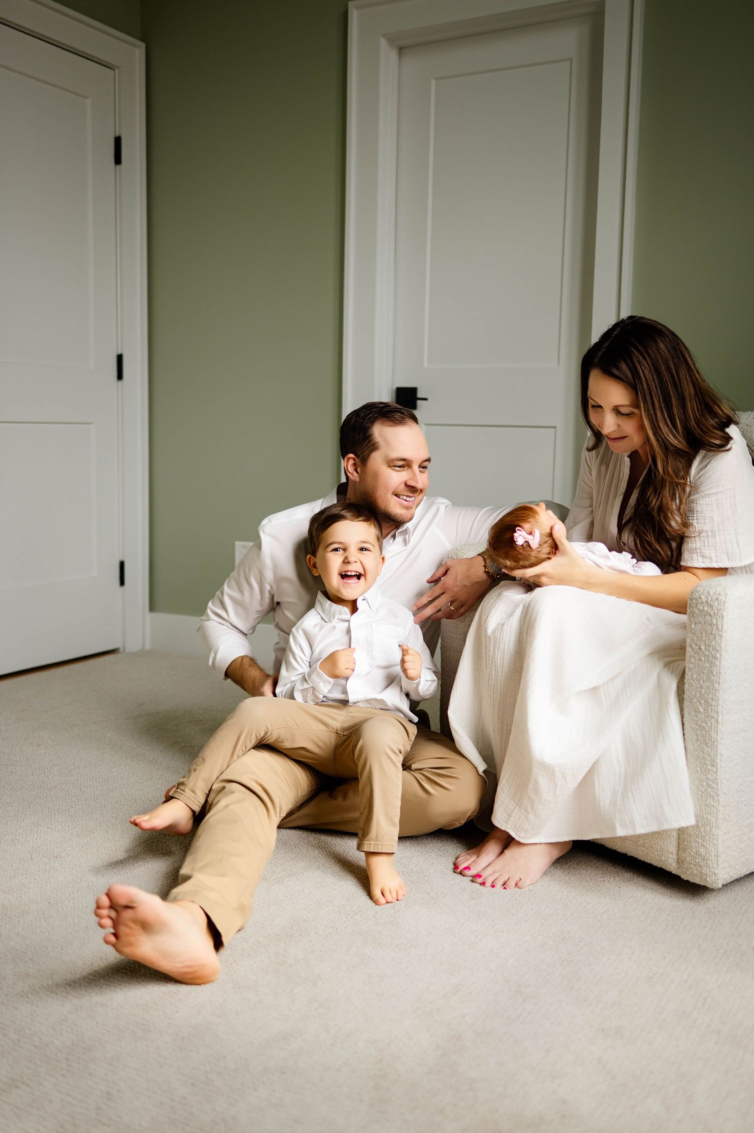 Family relaxes together on white carpet against sage green wall with matching white doors during indoor newborn photography Apex