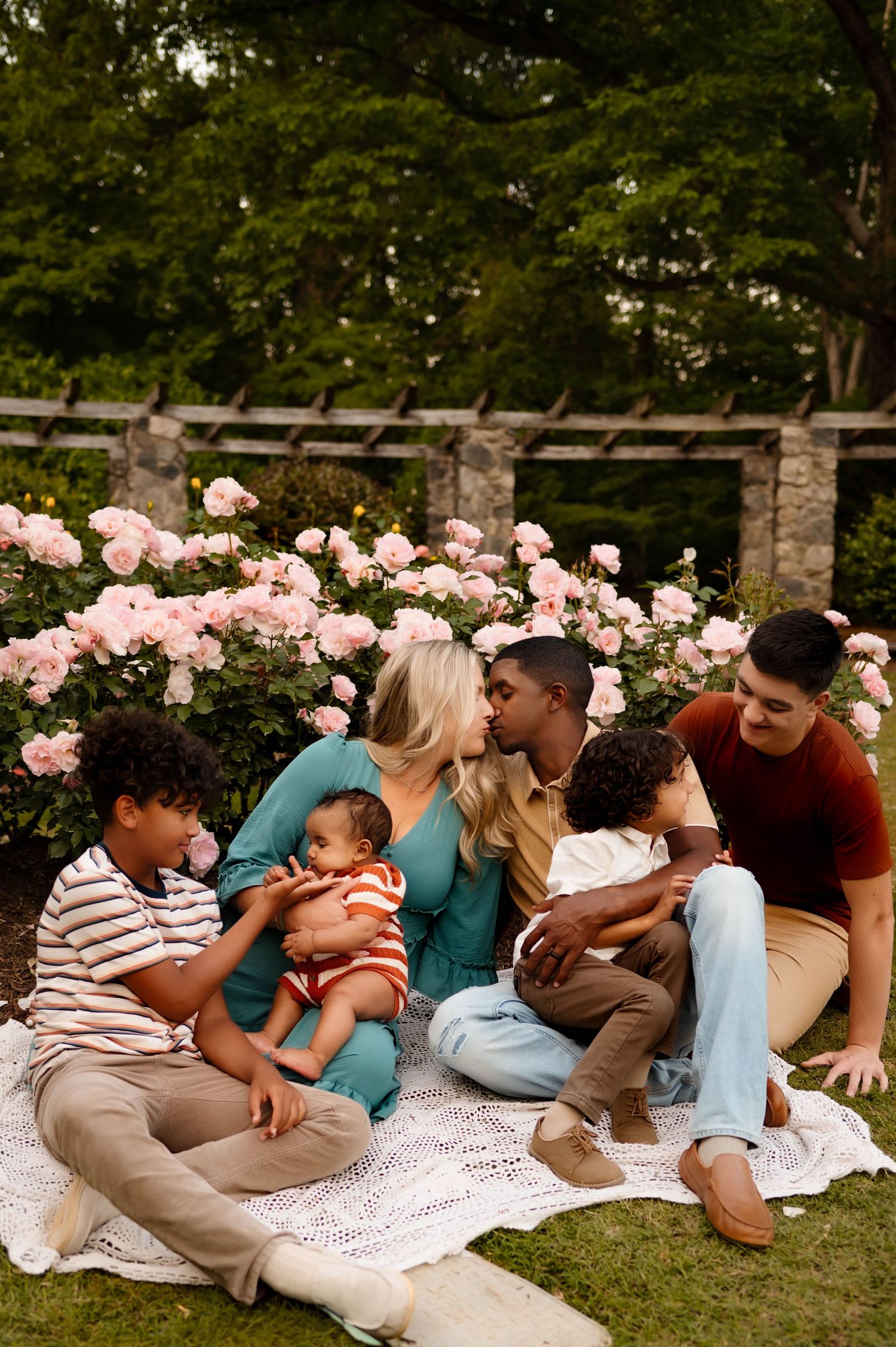 Family enjoying time together on blanket in garden with blooming roses while being photographed by family photographer in raleigh