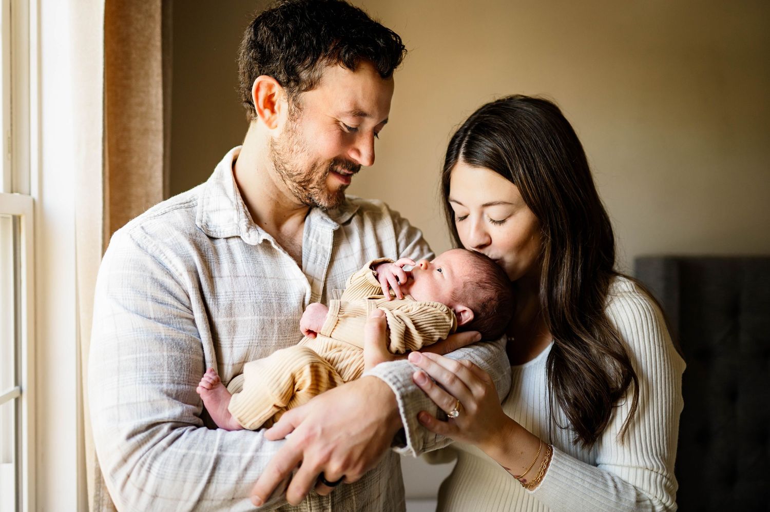 Parents smile lovingly while holding their newborn baby in a sunlit room near a window with newborn photographer wake forest