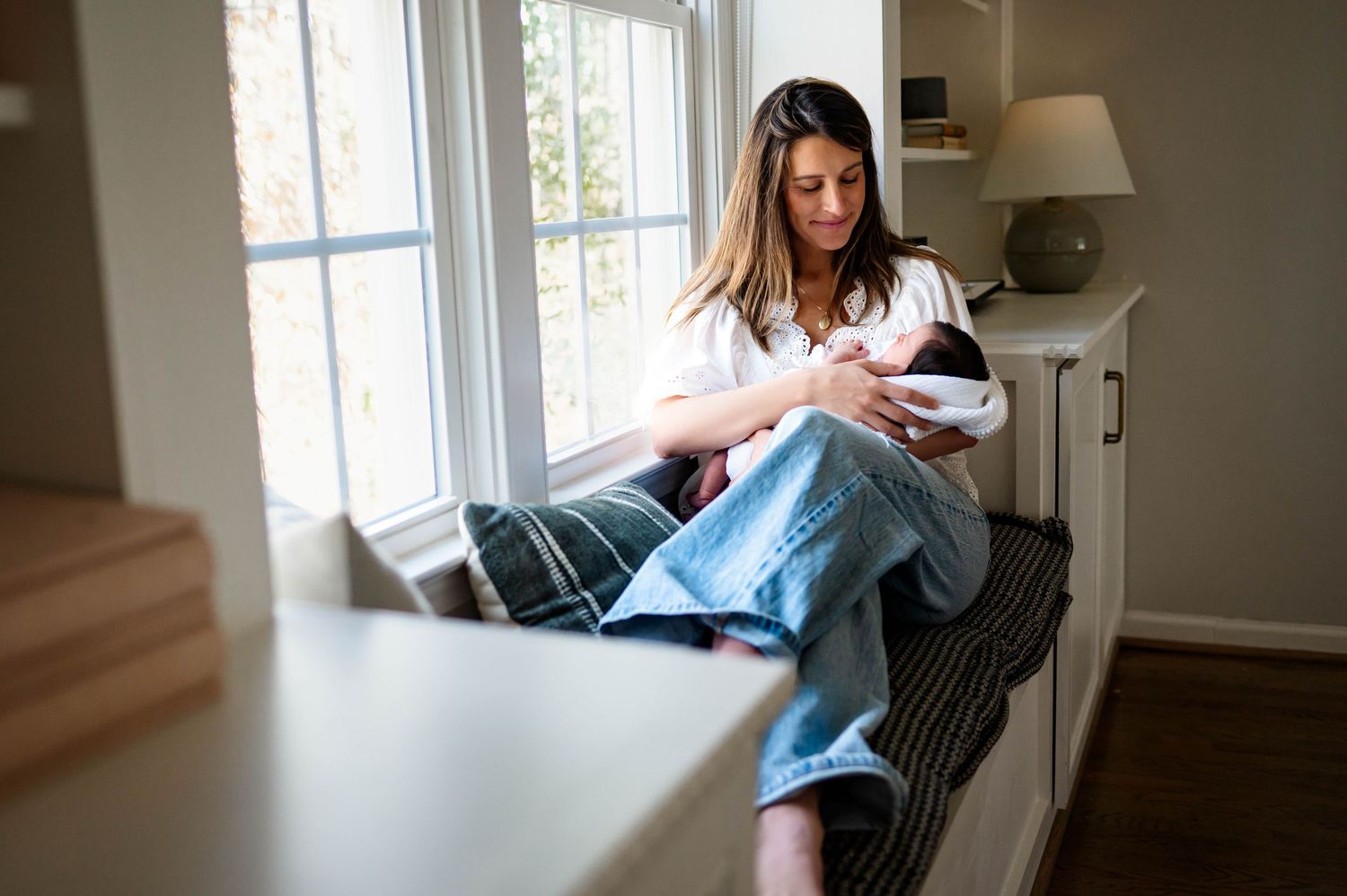 Mother dressed in white and jeans sits on a bench by a window with the best raleigh newborn photographer