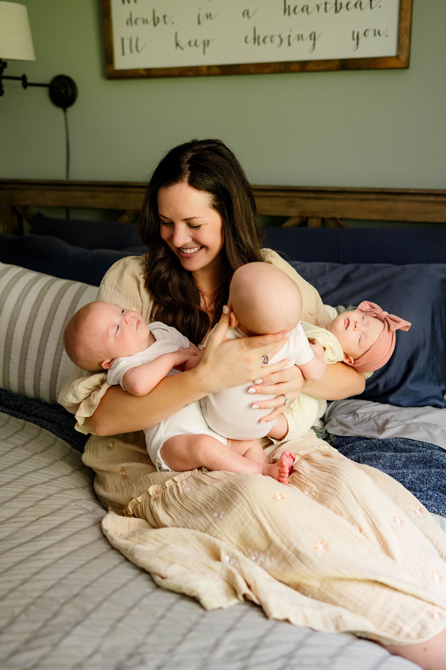 A mother in casual attire sits on a bed holding her three small infants in white onesies with raleigh newborn photographer