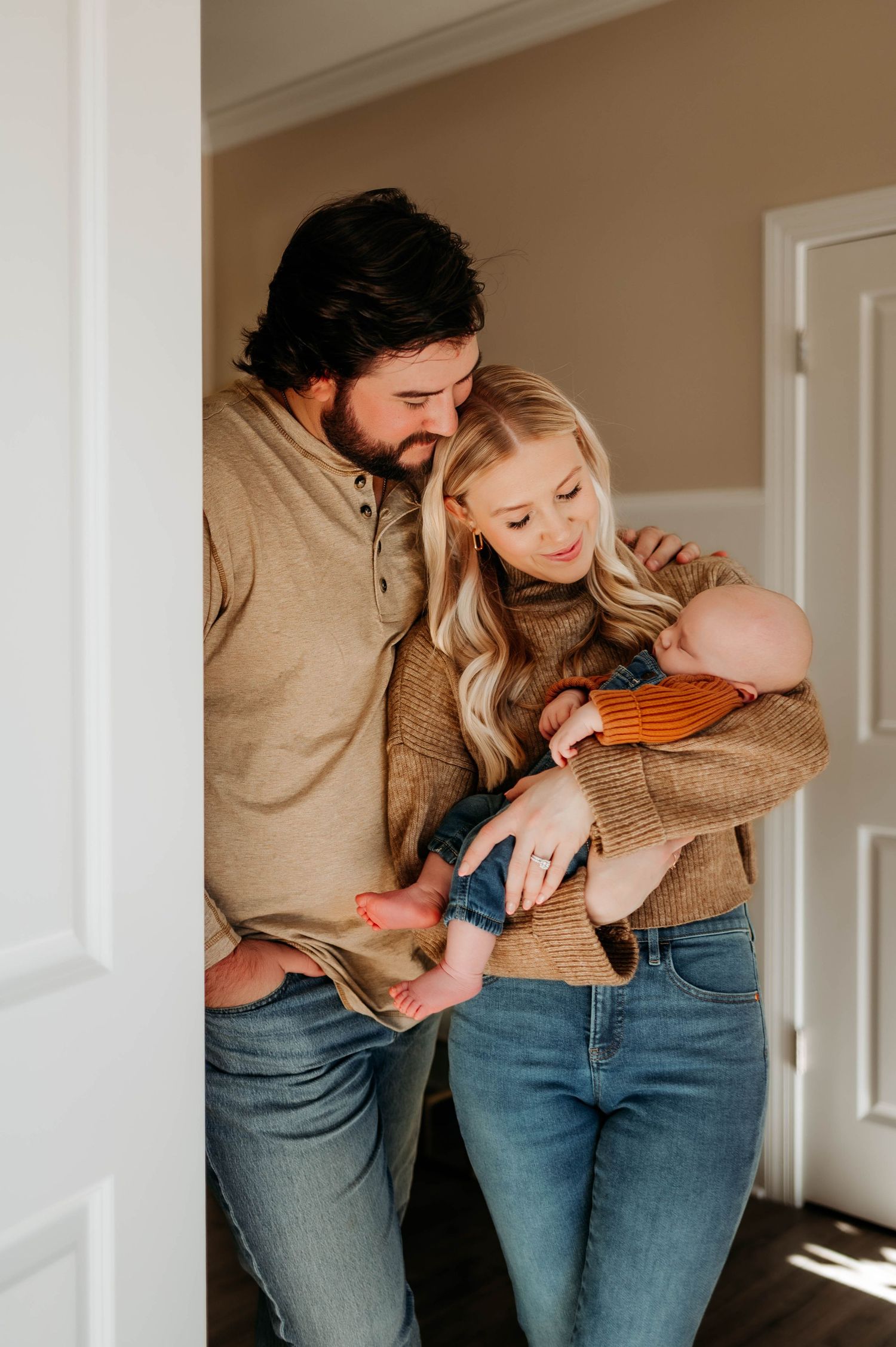 A couple in matching beige sweaters and jeans share a tender moment with their baby in a sunlit doorway at home during newborn photoshoot