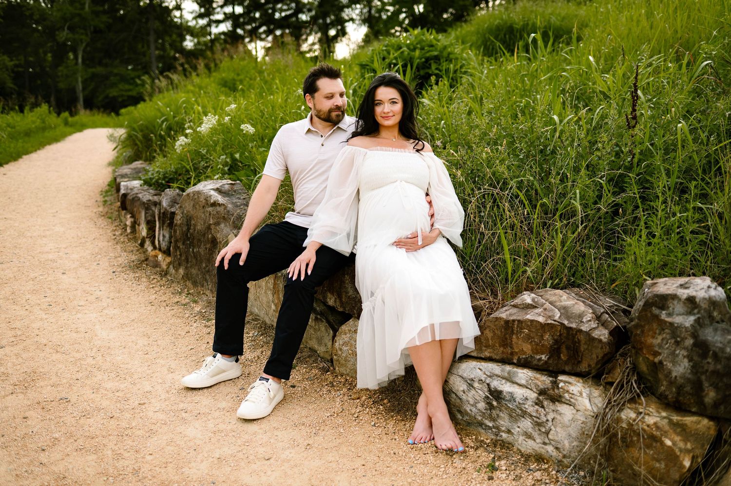 Couple sits together on stone wall along winding gravel path surrounded by lush green summer grass with apex maternity photographer