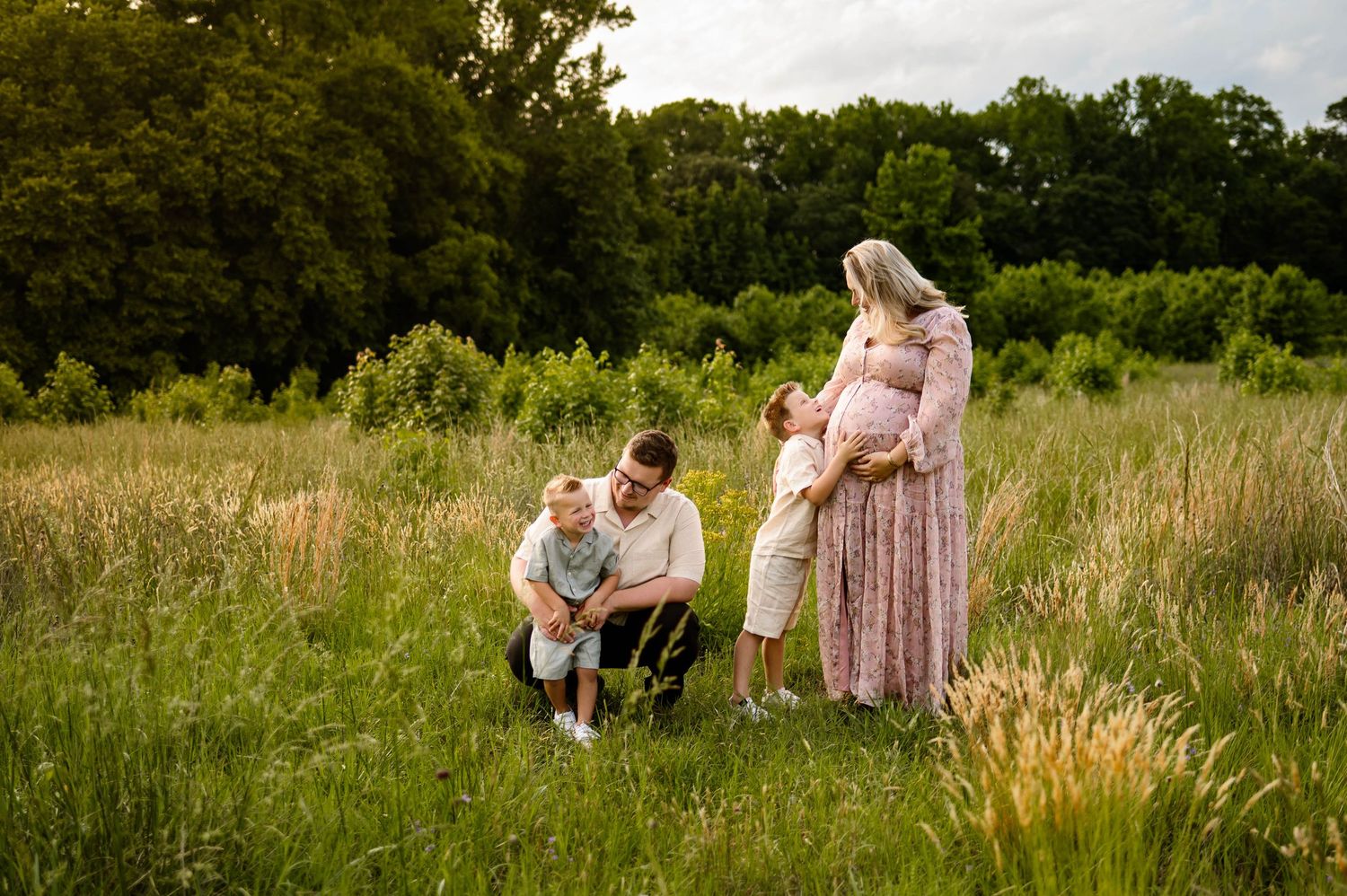 A family shares a tender moment together in a sunlit meadow with tall grass and trees in the background with raleigh maternity photographer