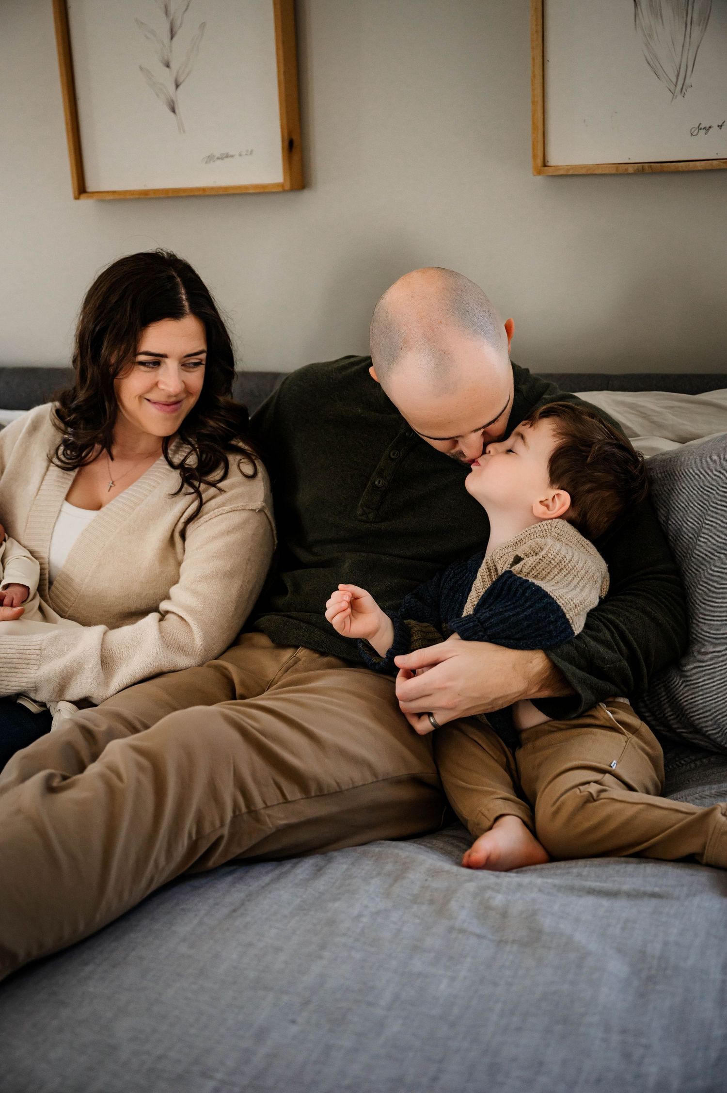 A family shares a cozy moment together while relaxing on a gray couch beneath framed artwork during newborn photoshoot in Raleigh, NC