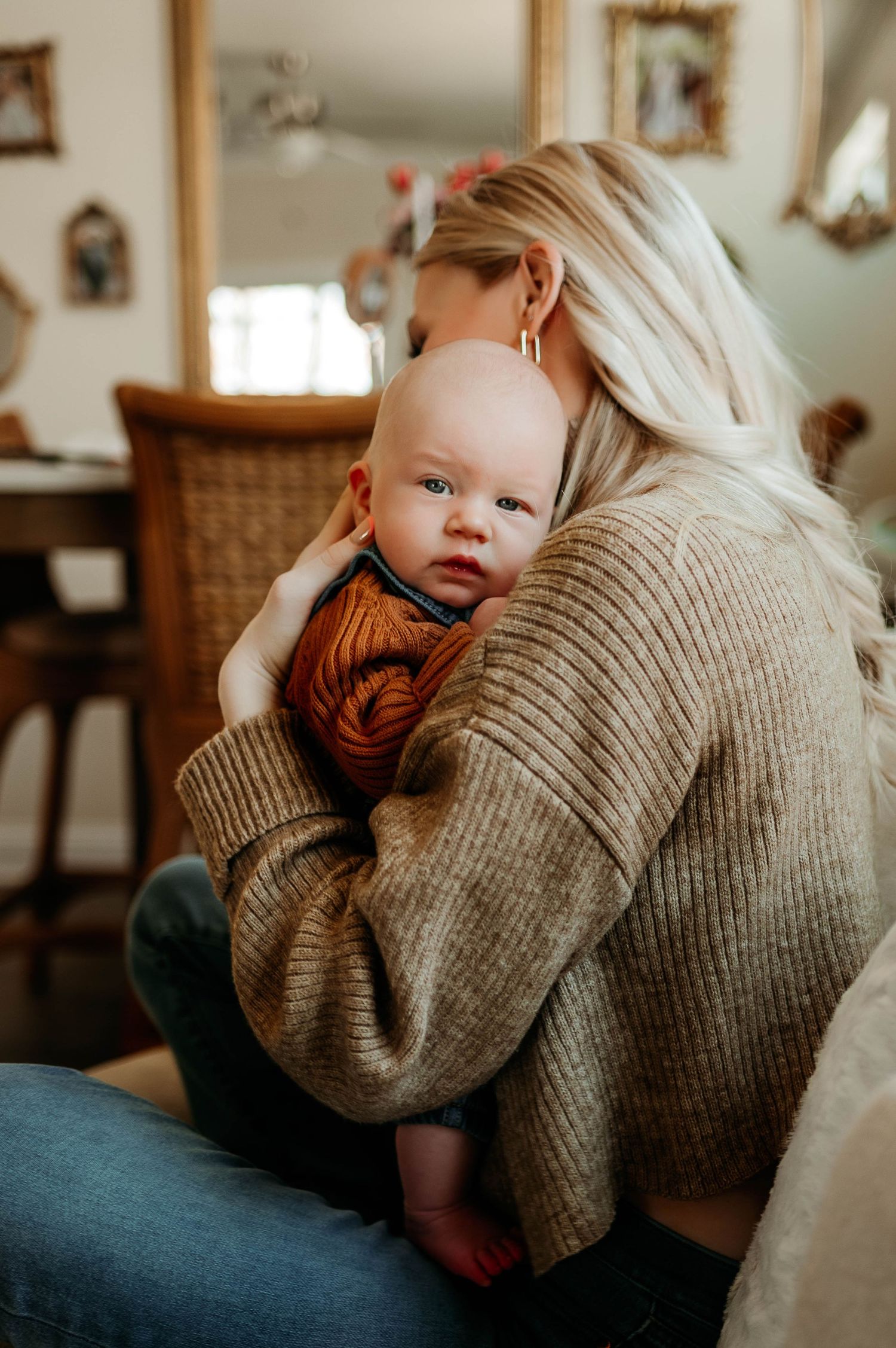 Baby in rust-colored top rests against mother wearing beige sweater in cozy home setting during newborn photography session raleigh nc