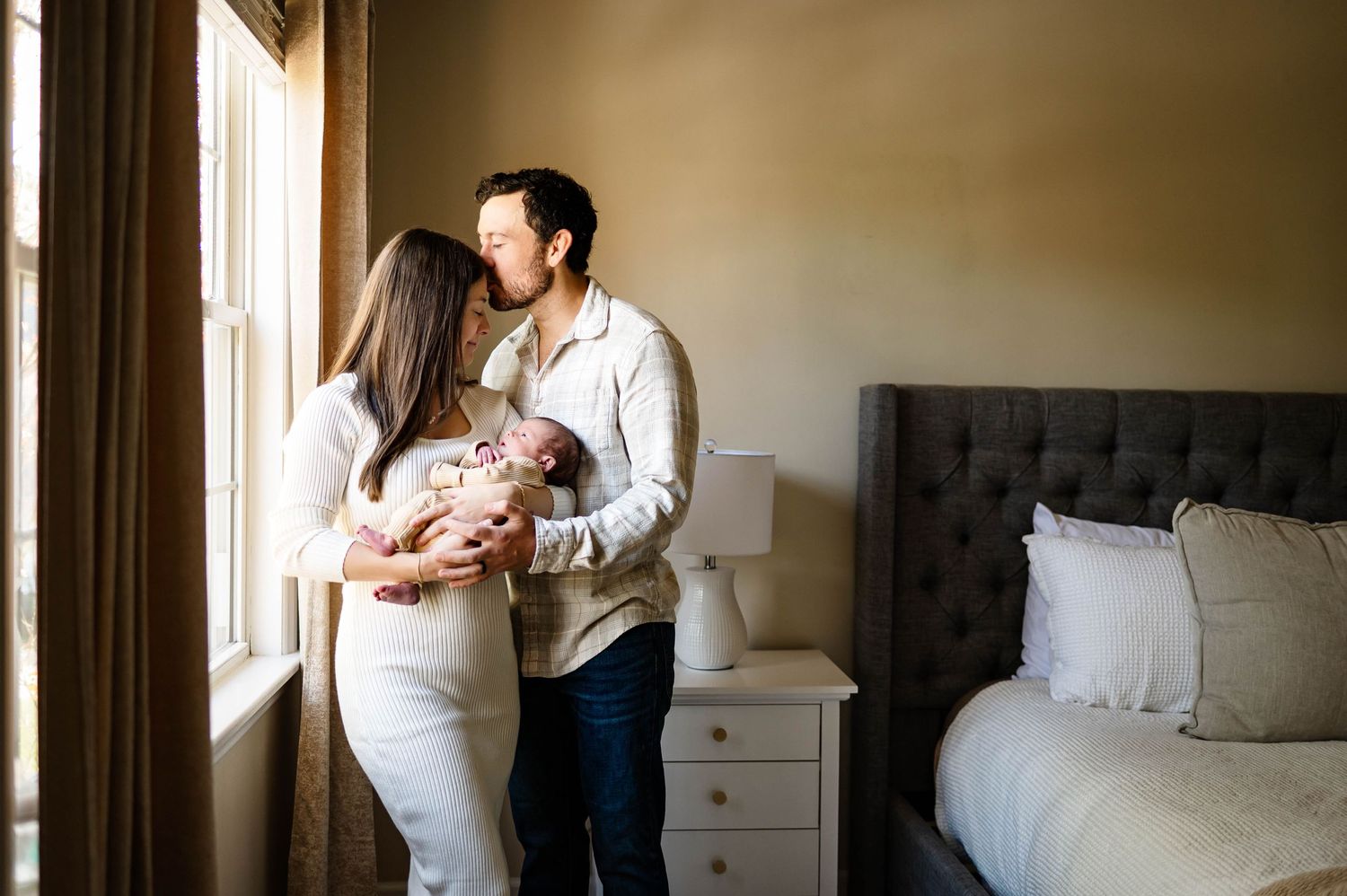 Couple in white and beige clothing sharing a caring moment by a window with wake forest newborn photographer