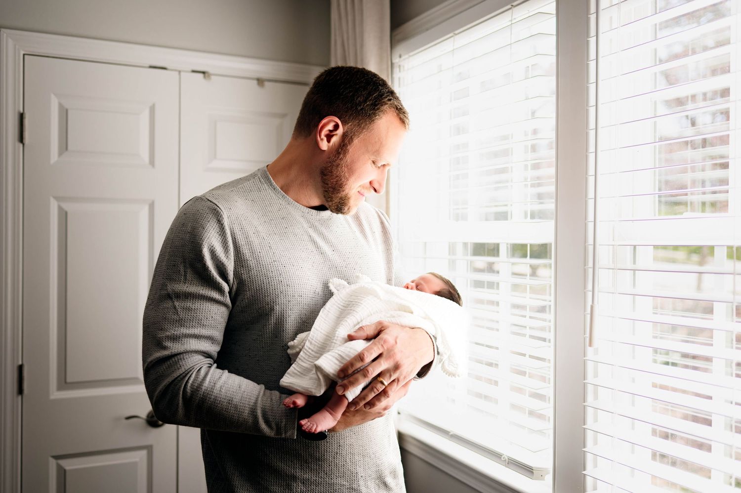 Father wearing a gray sweater holds a swaddled newborn baby during intimate newborn photoshoot with raleigh newborn photographer