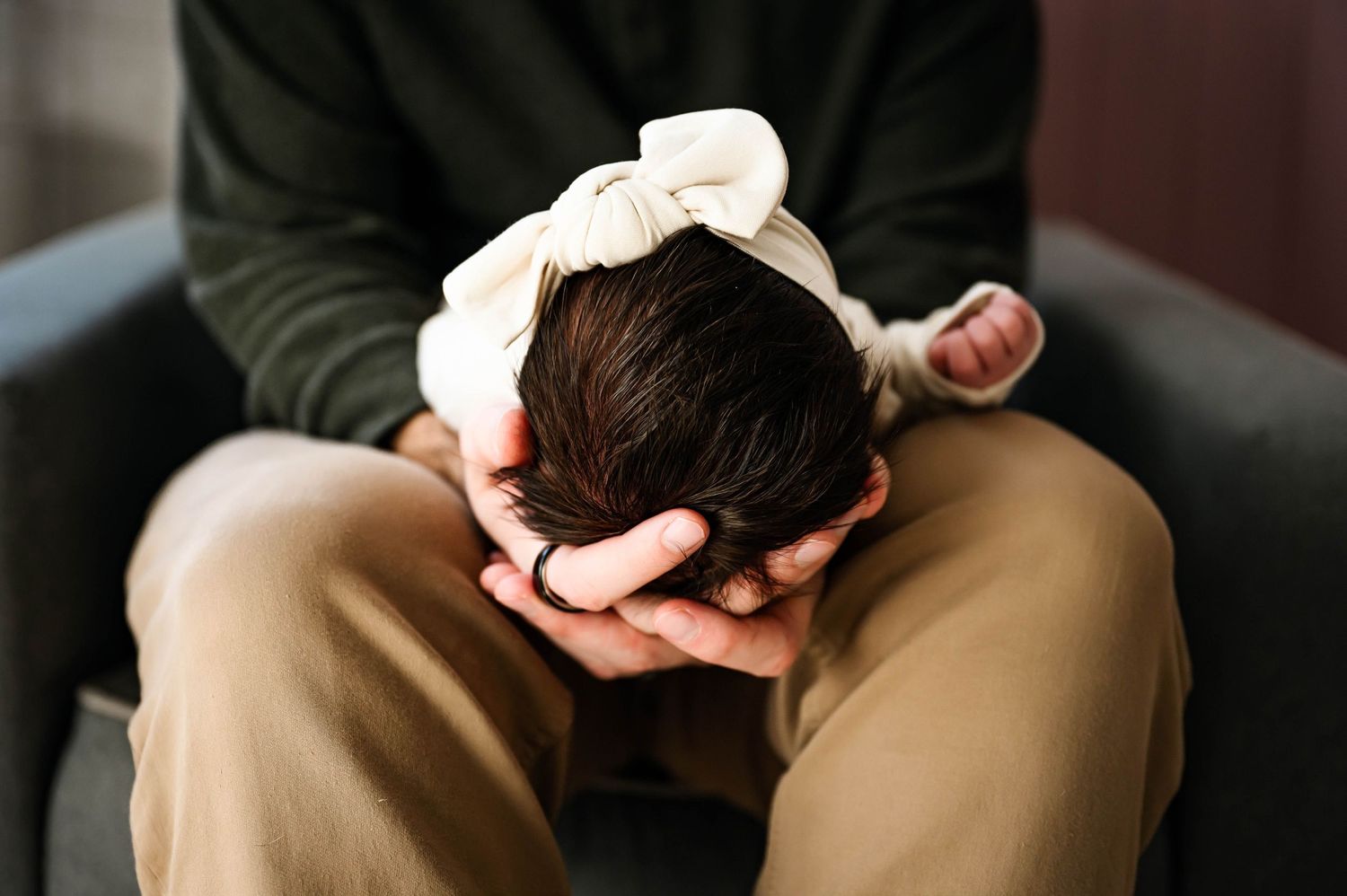 A newborn infant with dark hair rests in father's gentle hands while wearing a white bow headband in soft image by Emma Branch Photography