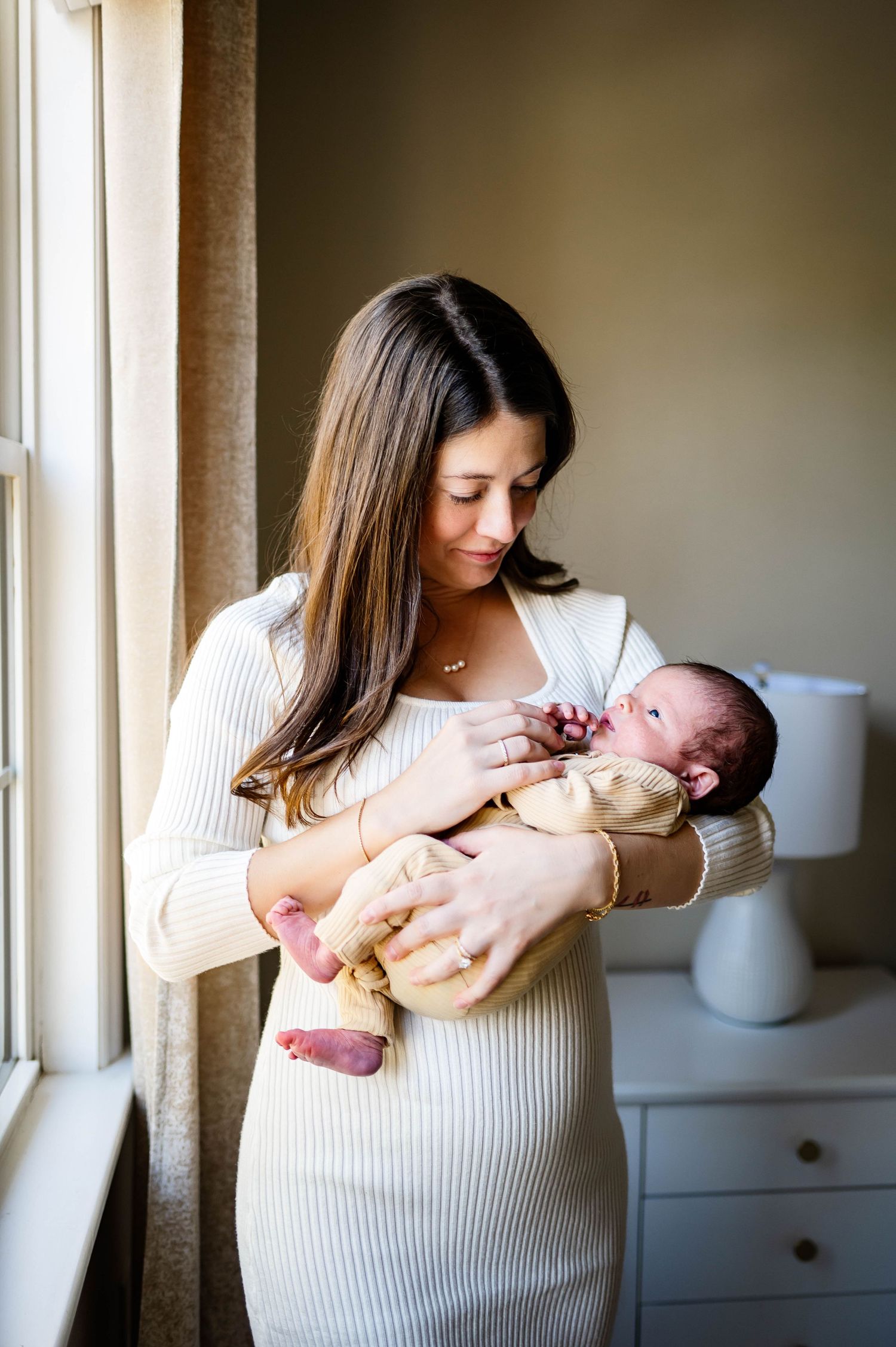 A tender moment by a window as a mother in a white ribbed dress cradles her newborn baby with wake forest newborn photographer