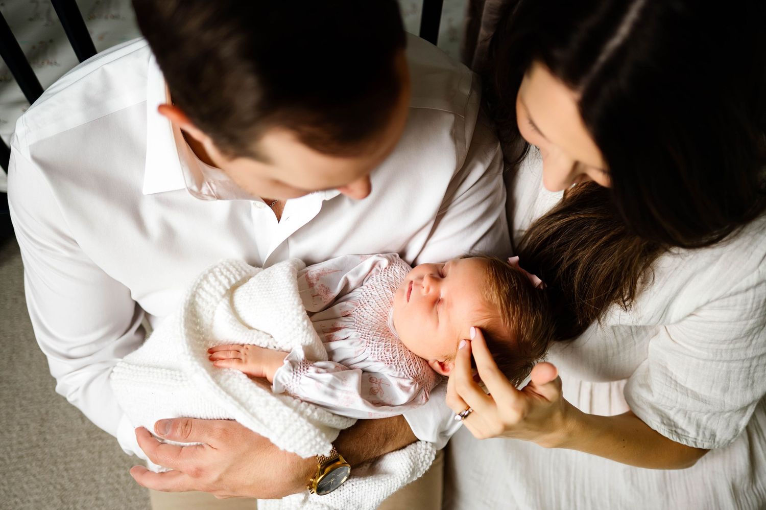 A newborn baby in white lace clothing lies between parents in white shirts while smiling and laughing with newborn photographer raleigh