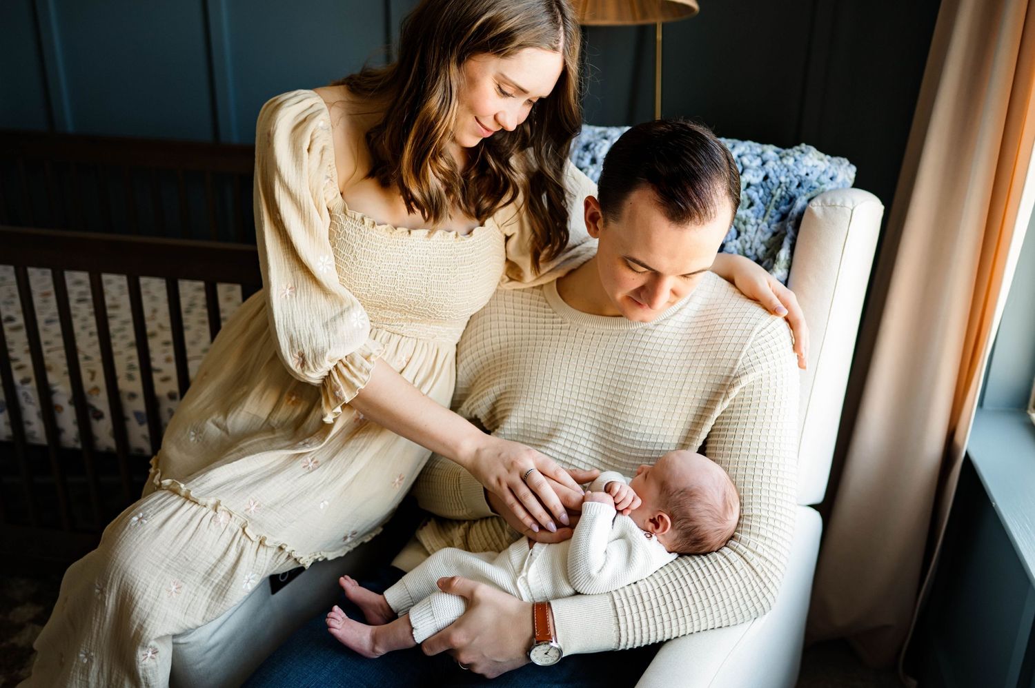 A couple in matching neutral sweaters cradles their newborn baby near a dark wooden crib in a nursery with best apex family photographer