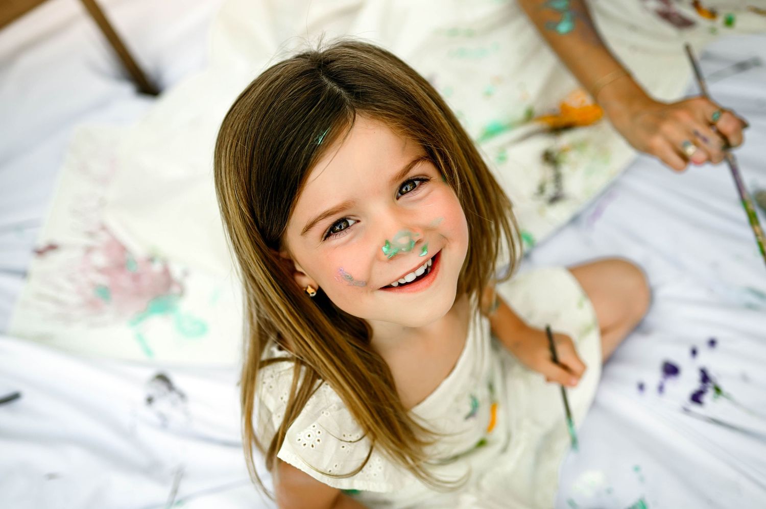 Little girl smiles at camera while sitting on paint-splattered white canvas during a family photography shoot with cary family photographer
