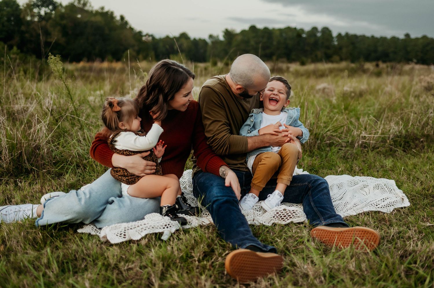 Family laughs together on blanket in field during golden hour outdoor photo session with wake forest family photographer