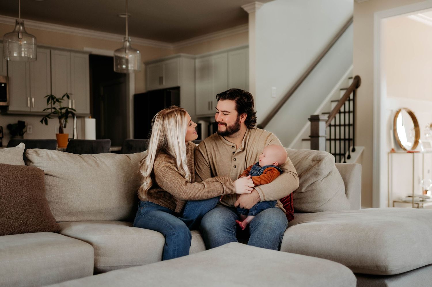 A couple sitting on a gray sectional couch in their living room share a laugh while relaxing at home with apex newborn photographer