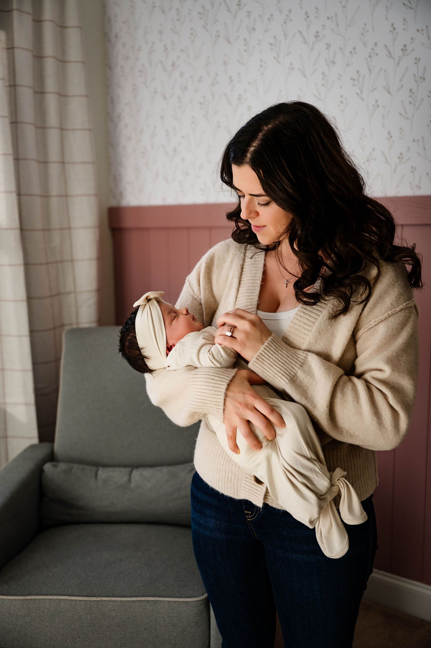 Mother in beige sweater tenderly cradles newborn baby while standing beside gray chair during shoot with Emma Branch Photography