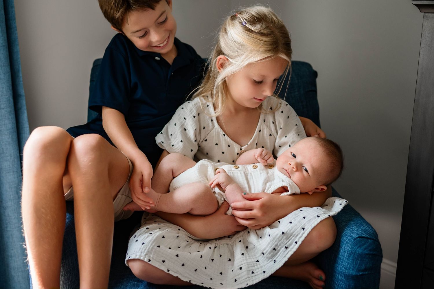 Siblings sit together on blue furniture while carefully holding and looking at their baby brother with newborn photographer apex