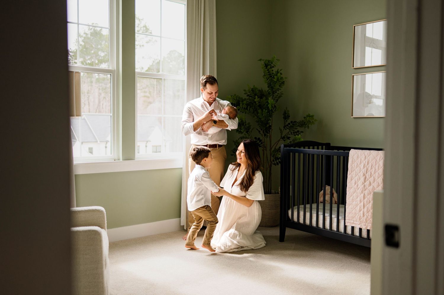 A family sits together in a minimalist nursery with green walls, natural light streaming through windows with newborn photographer near me