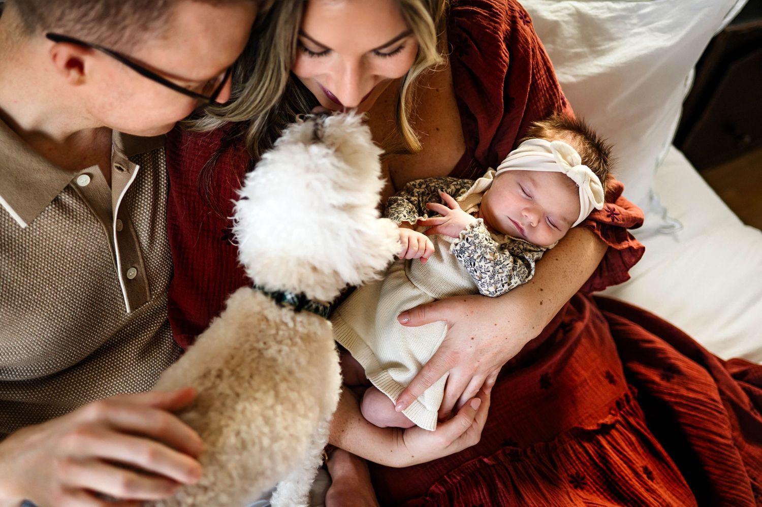 A small white dog lovingly interacts with baby held by parents on a cozy bed during newborn photo session in durham nc