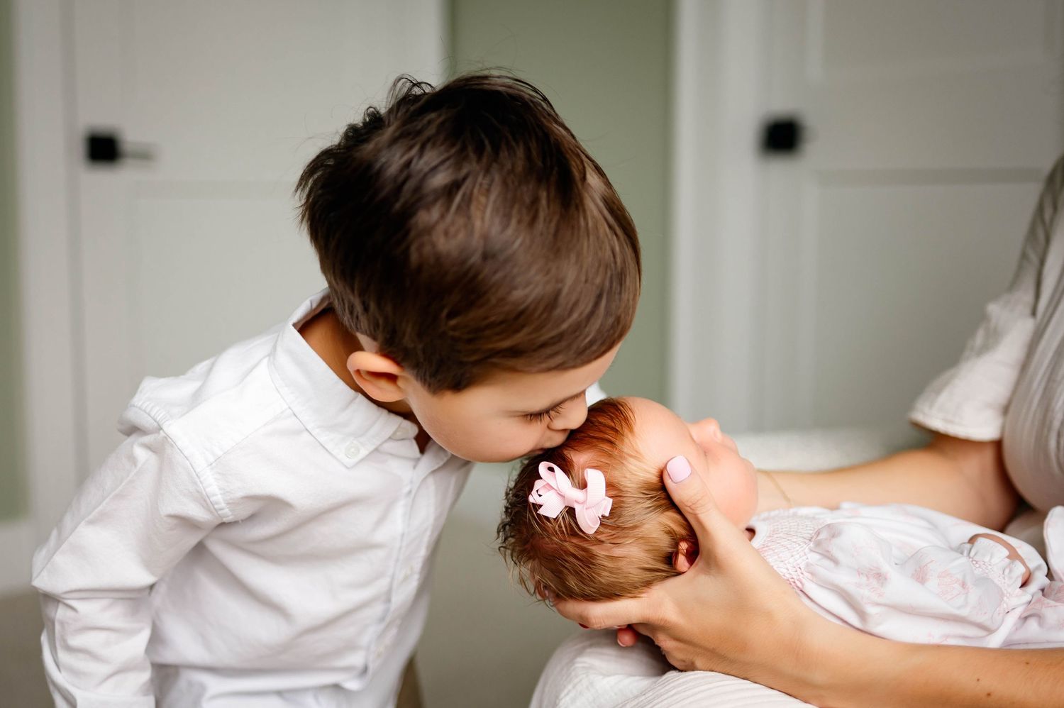 A child in a white shirt gently kisses newborn sister swaddled in white during intimate newborn photography session with Durham photographer