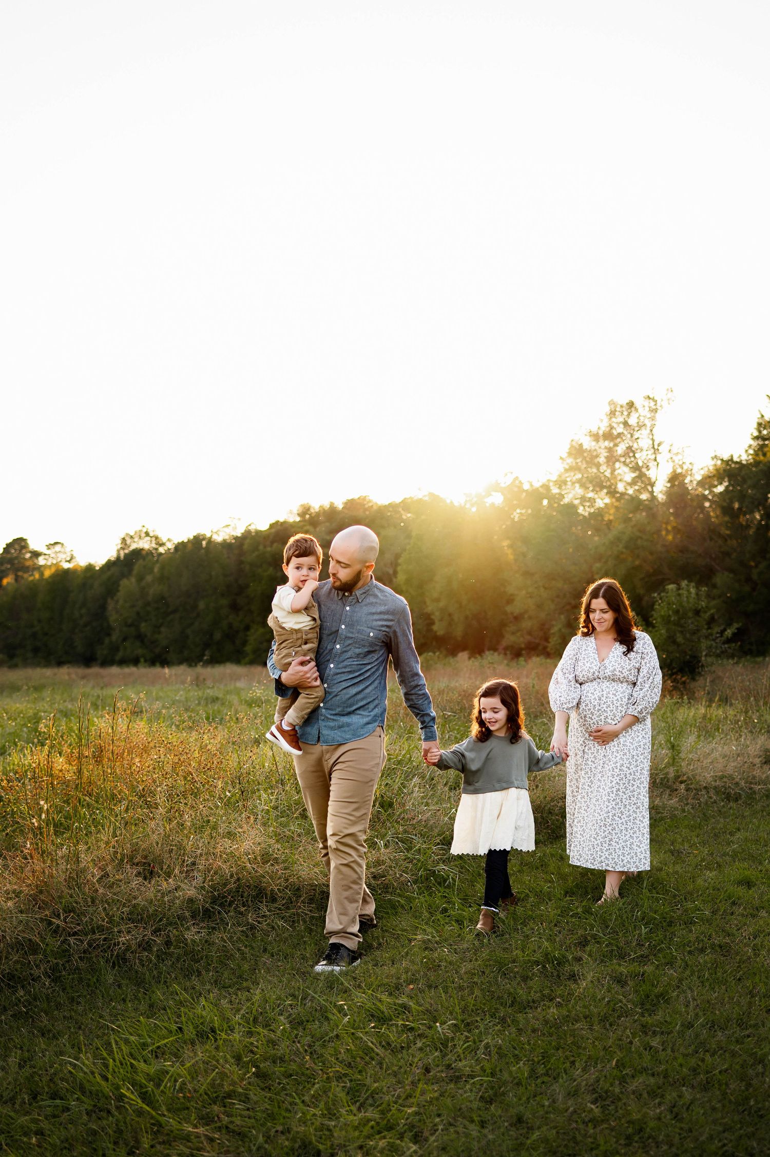 Family walks together through a sunlit meadow at golden hour during a warm summer evening during a family photography session in raleigh nc