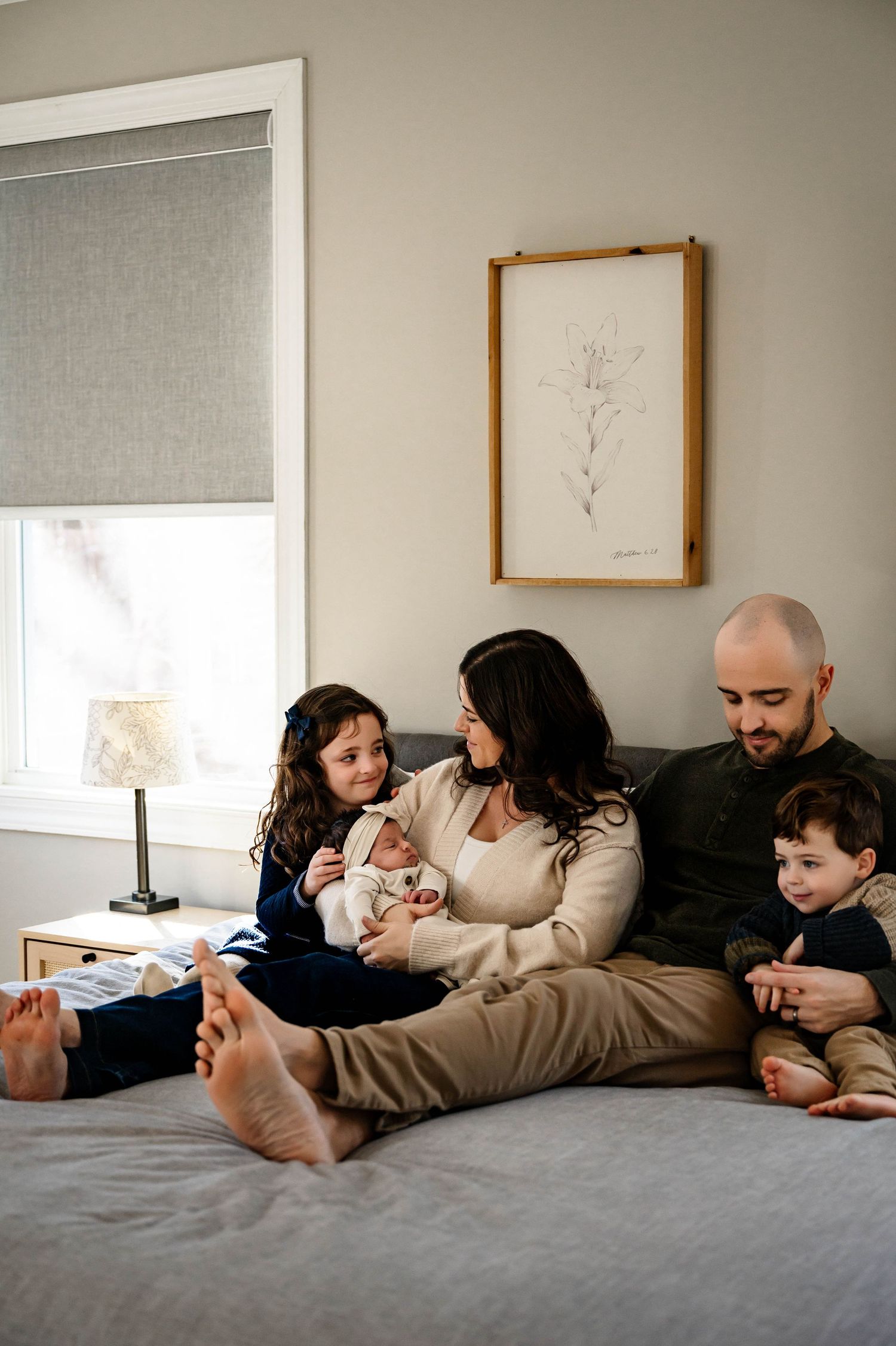 Family relaxes together on gray bed while spending quality time at home for durham newborn photographer