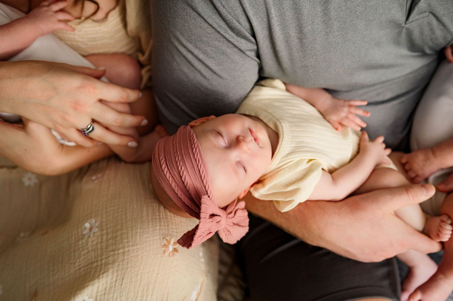 A sleeping newborn baby wearing a pink headband lies peacefully in their parents' arms with durham newborn photographer in home