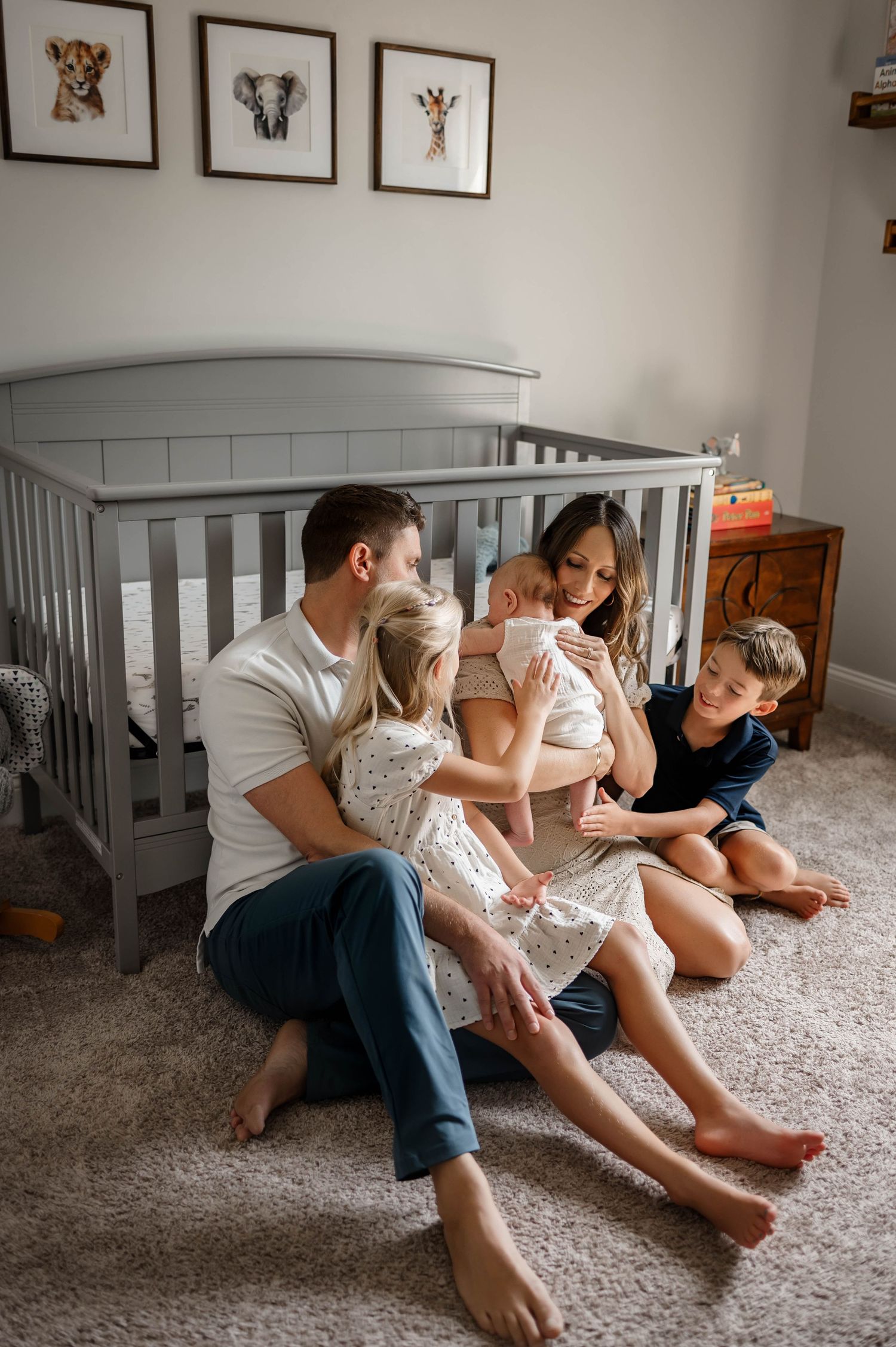 In acozy nursery family sits on carpet in front of a grey crib, with animal artwork displayed during newborn photography session raleigh