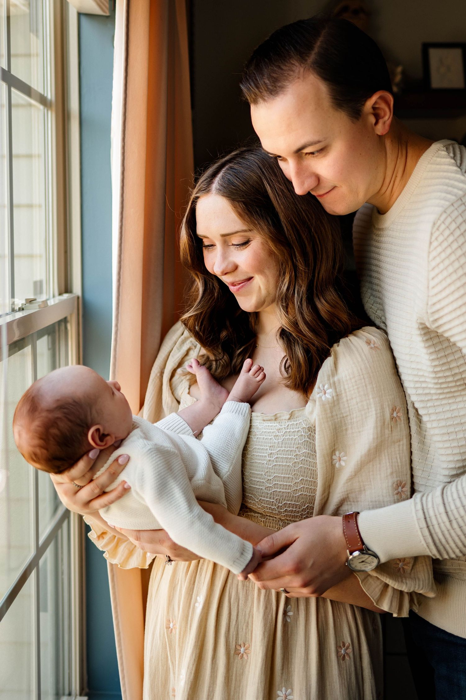 A tender family moment by a window as loving parents gaze adoringly at their newborn baby with raleigh newborn photographer