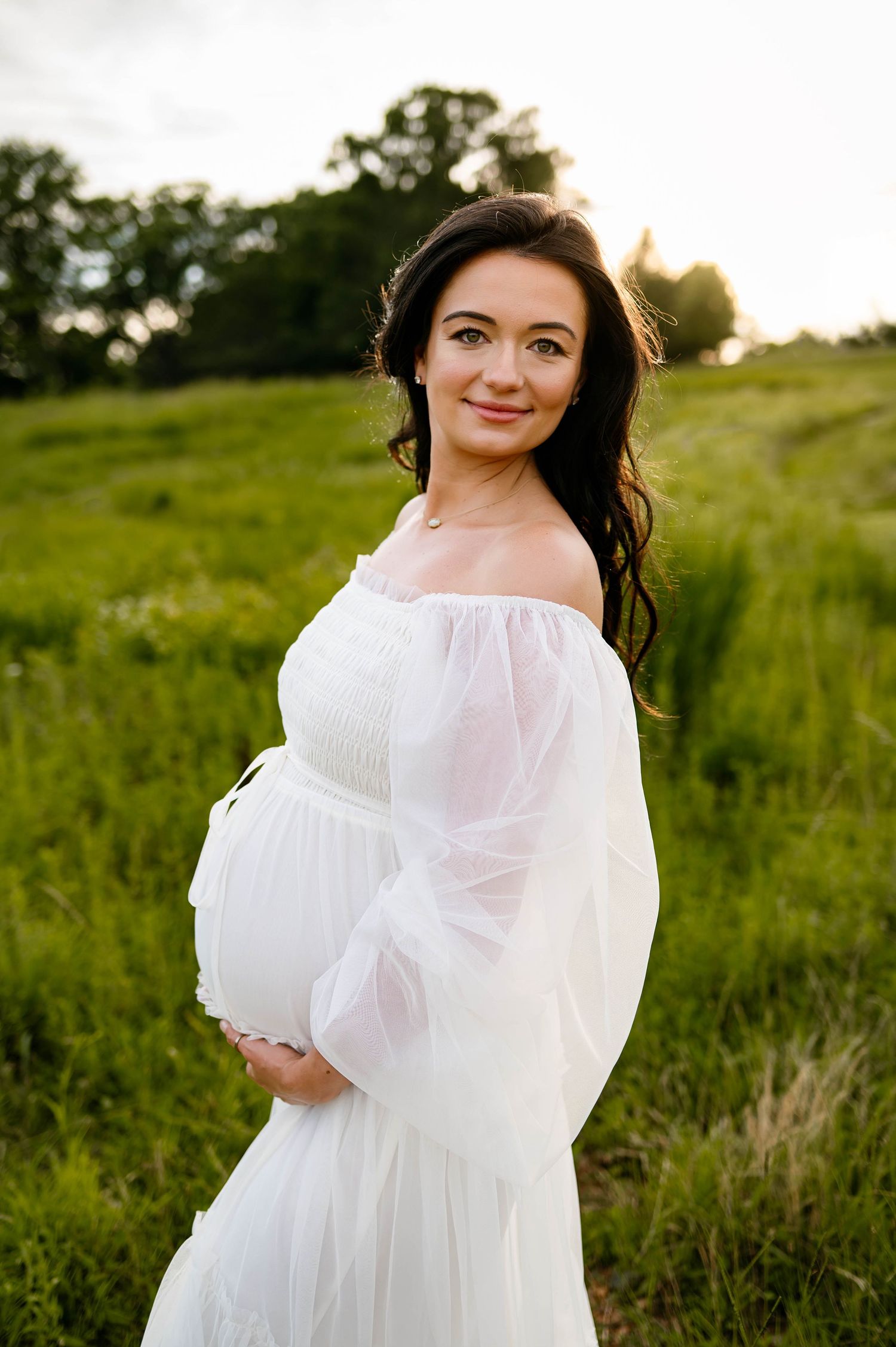 An expectant woman in a white flowy dress poses in a grassy field at sunset with wake forest maternity photographer