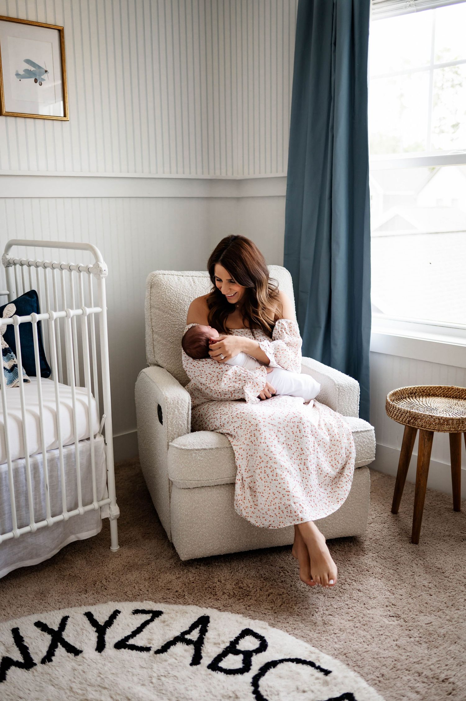 A nurturing moment in a cozy nursery with white crib, beige recliner, blue curtains and alphabet rug with raleigh newborn photographer